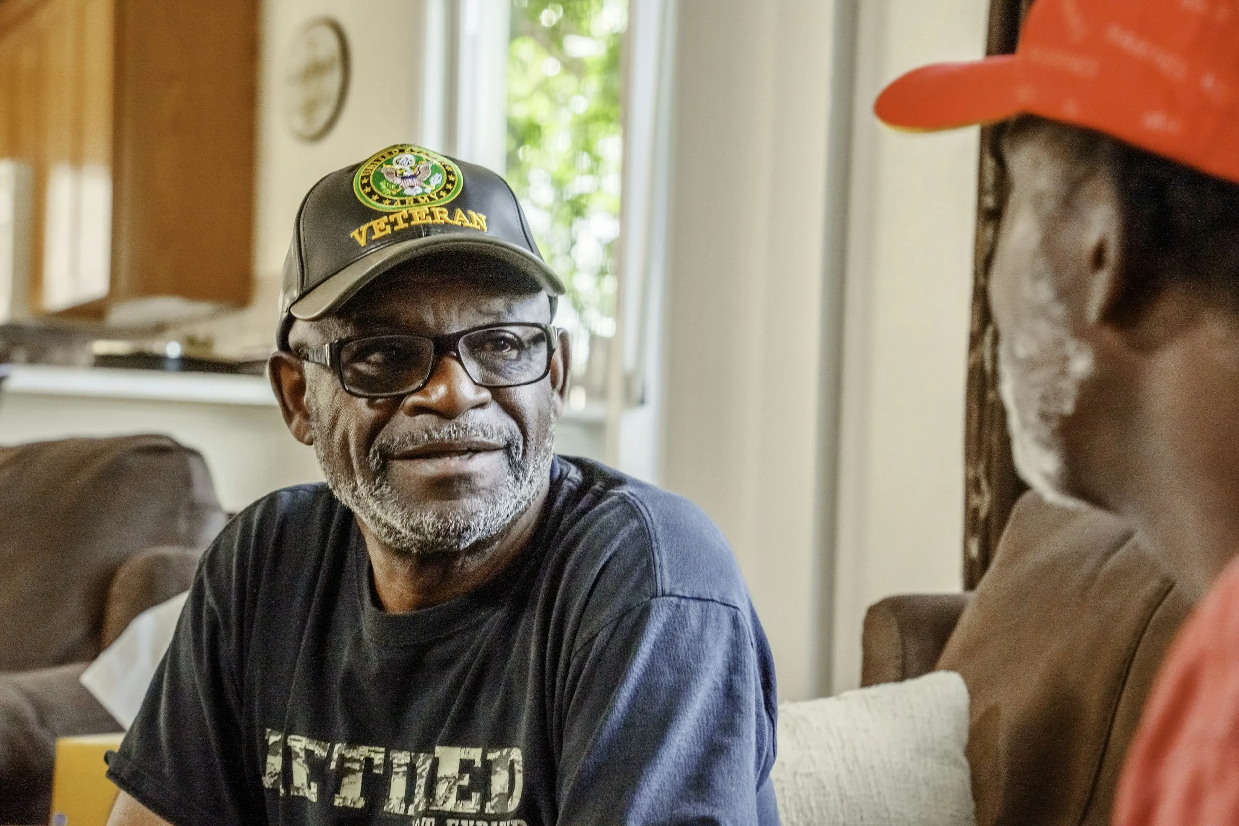 An elderly man with glasses, a gray beard, and a black hat with a veteran patch, sitting on a couch and smiling while talking to a person in a red hat.