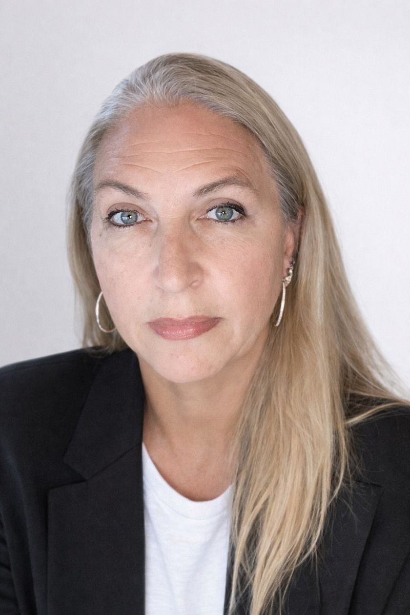 A woman with long blonde hair, blue eyes, wearing a black blazer and a white shirt, looking at the camera against a plain white background.