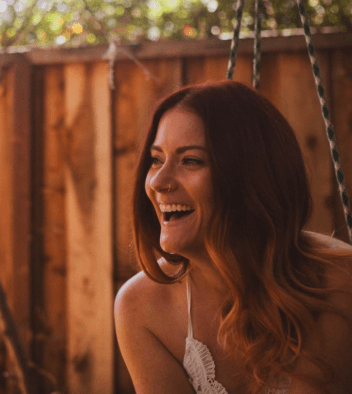 L.E. Sells, author of Kindred (Blood Rite) and Survive (From the Ashes). A redheaded woman smiling against a wooden fence backdrop