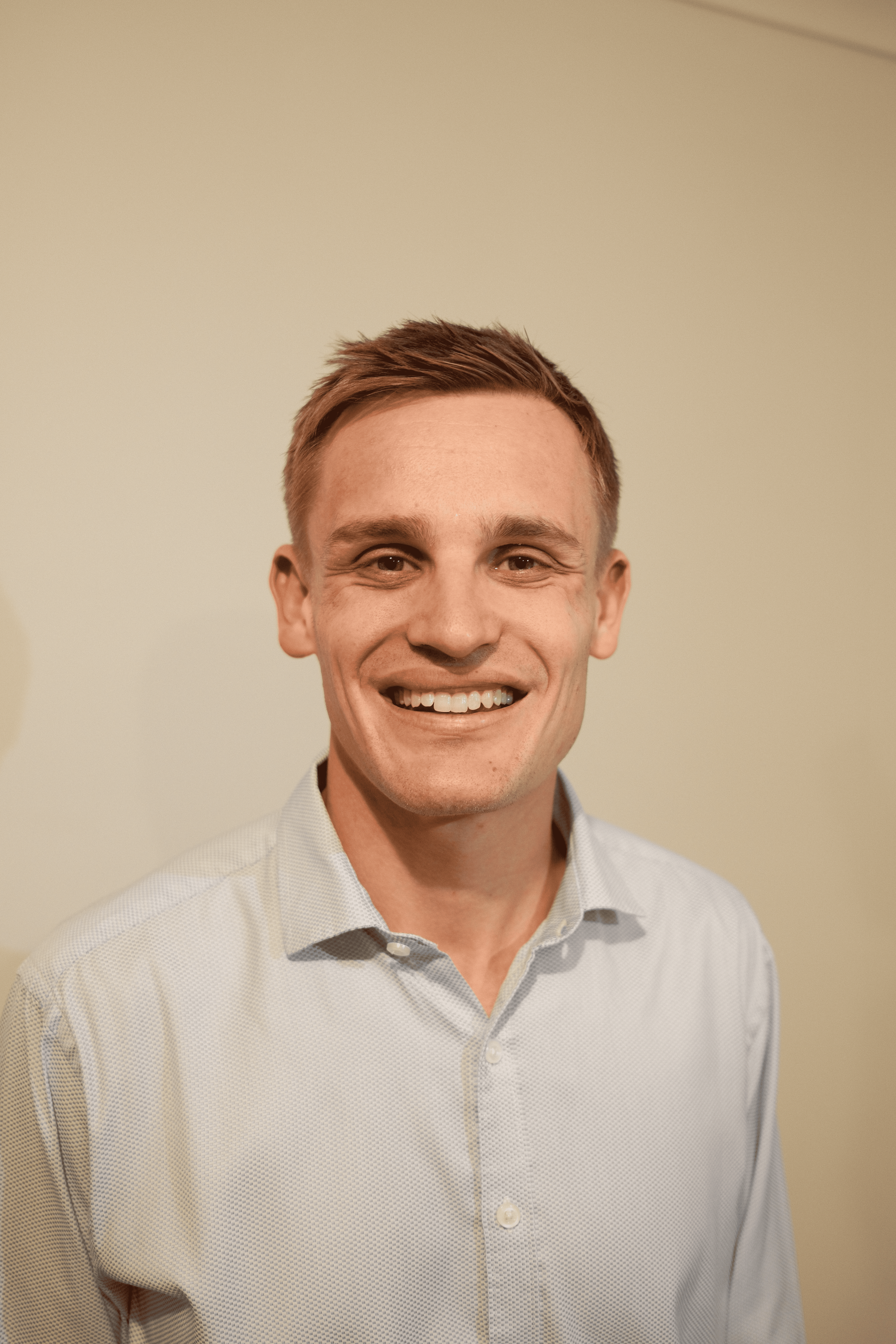 A smiling young man with short reddish-brown hair wearing a light-colored, collared shirt standing against a plain light-colored wall.