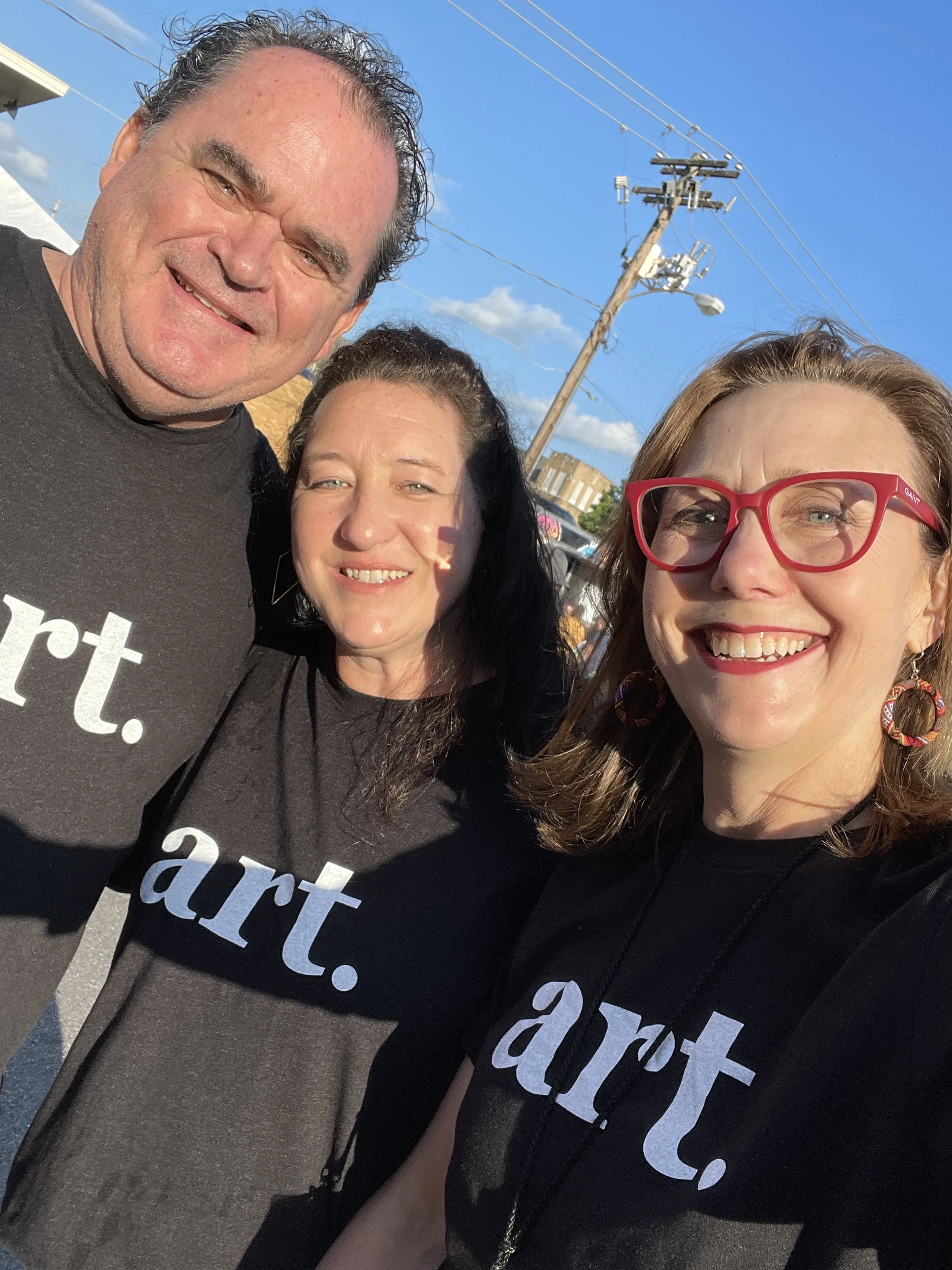 A group of three smiling people taking a selfie outdoors on a sunny day, with a power line and blue sky in the background.