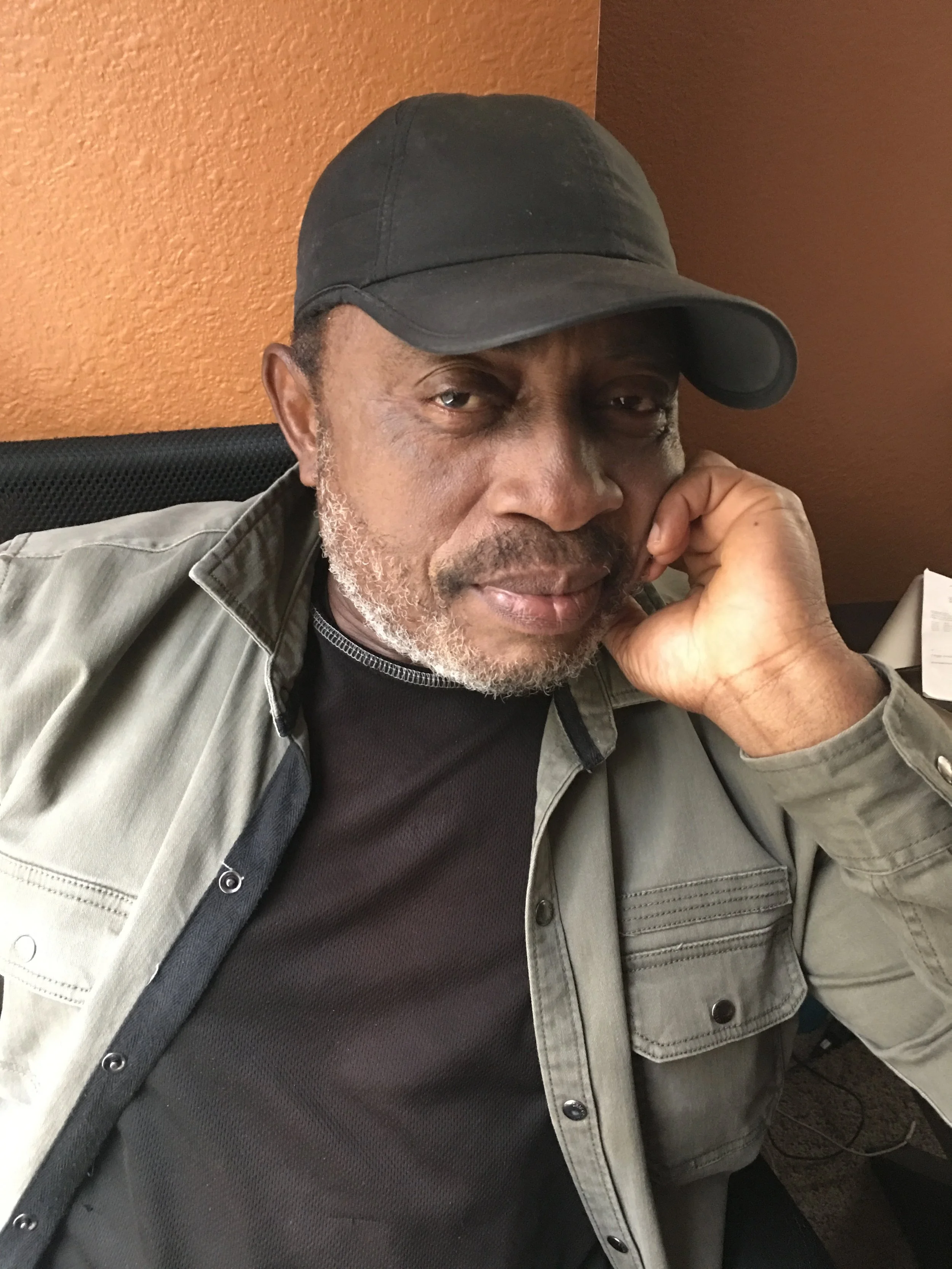 A man wearing a black cap, gray shirt, and black undershirt sits at a desk, resting his chin on his hand, in front of a burnt orange wall.