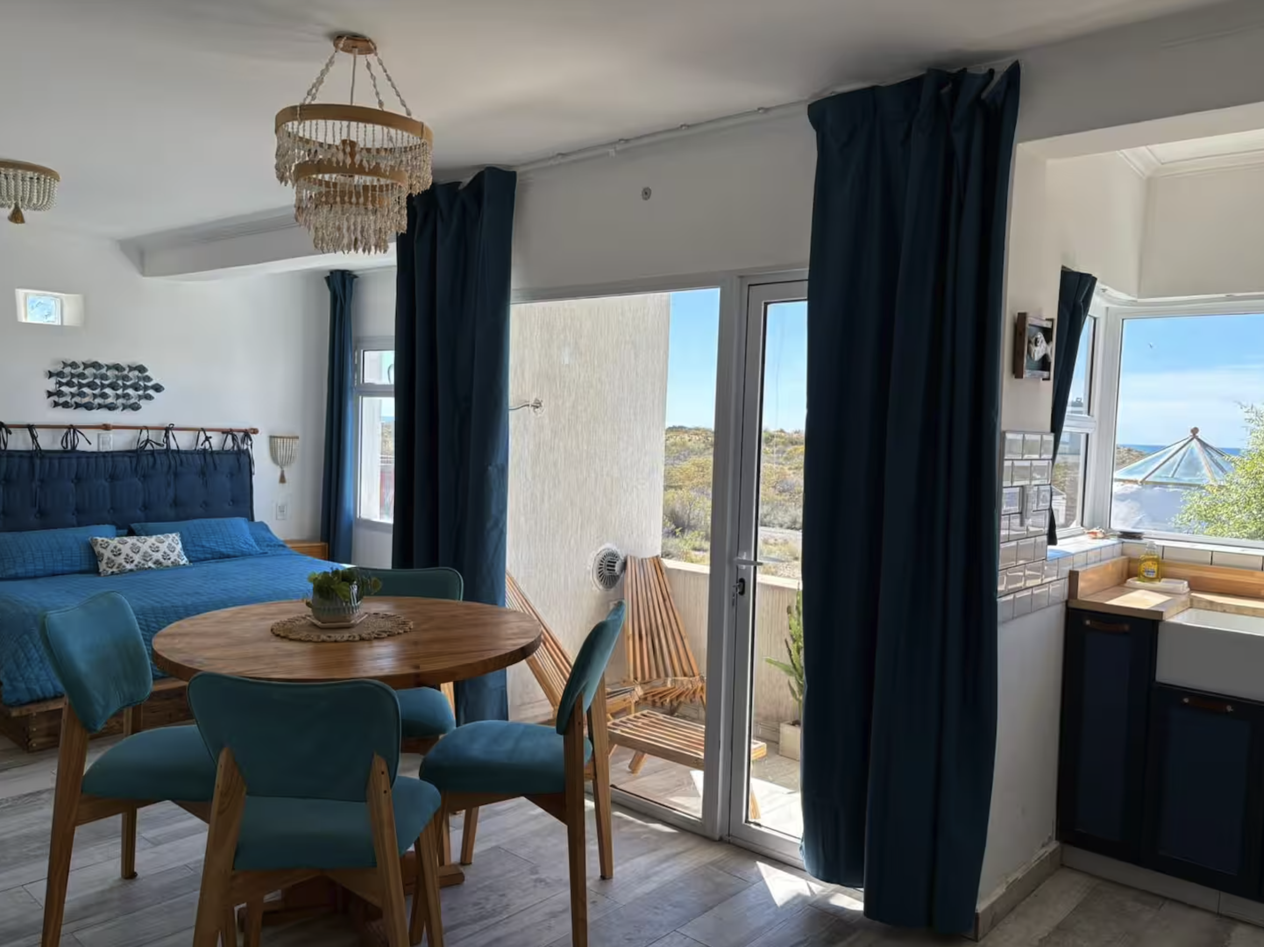 Interior view of a room with a blue bed, a round wooden dining table with four teal chairs, dark blue curtains, and sliding glass doors leading to a balcony with outdoor furniture and a landscape with hills and a blue sky visible outside.