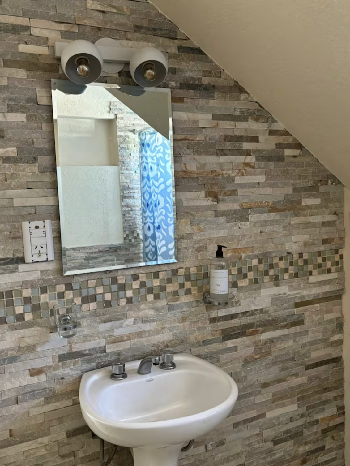 Bathroom sink with mirror and wall-mounted soap dispenser, set against a decorative stone wall.