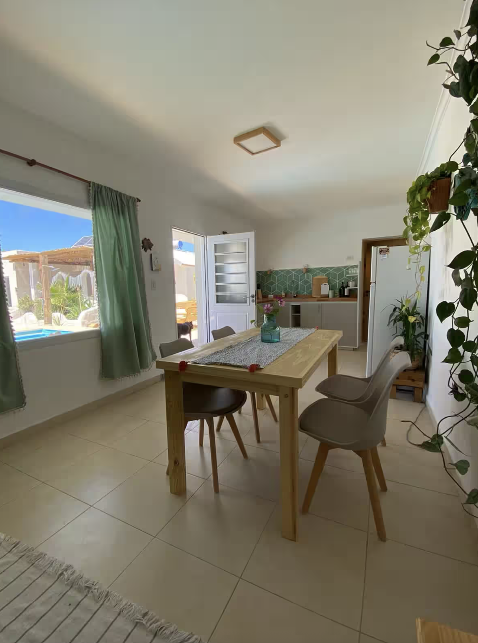 Dining room with a wooden table, four chairs, green curtains, potted plants, and a view of an outdoor patio.