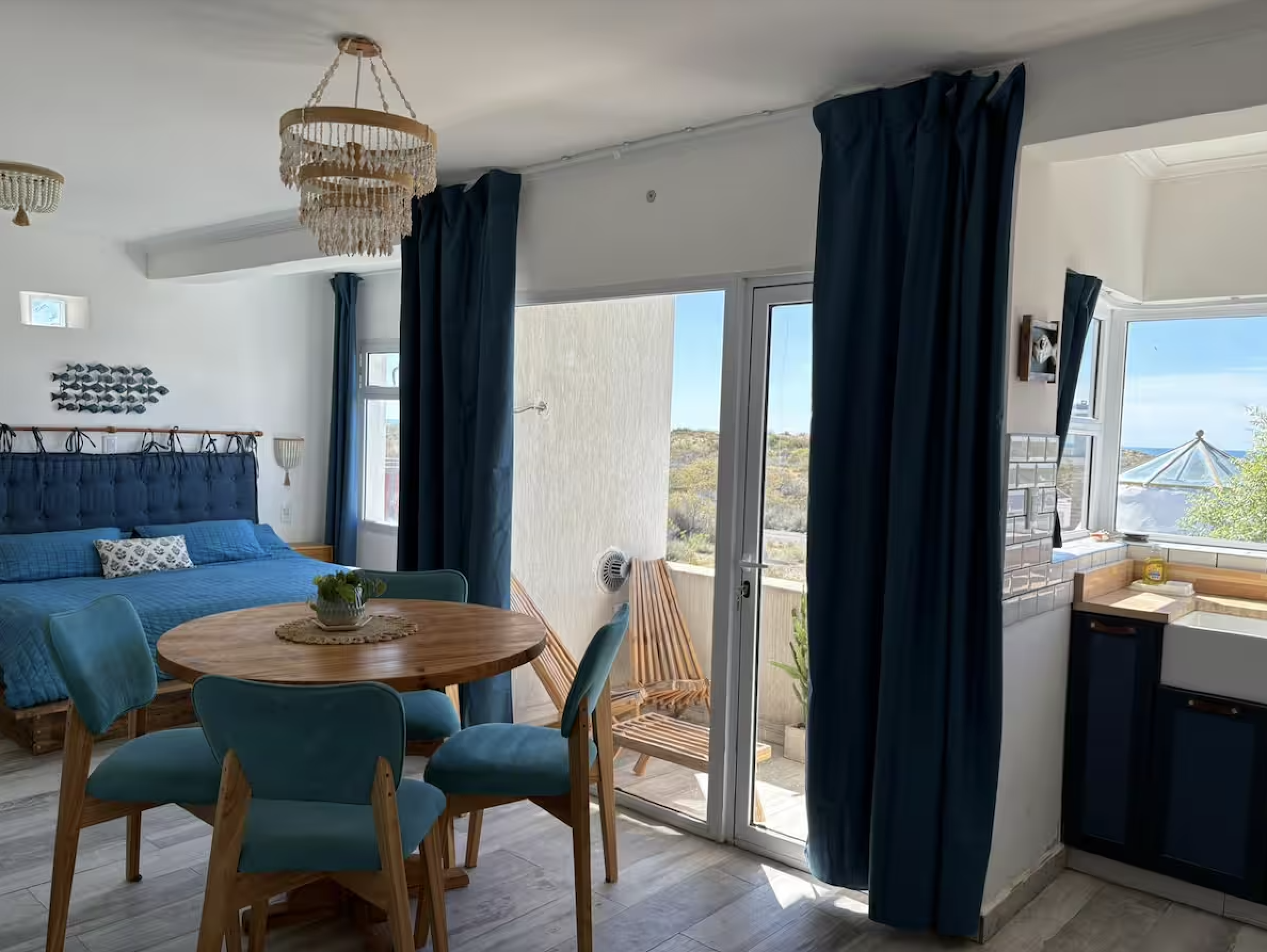 Coastal style living room with a blue bed, a round wooden dining table with teal chairs, and large windows with dark blue curtains showing an outdoor patio with wooden lounge chairs and a view of the ocean.