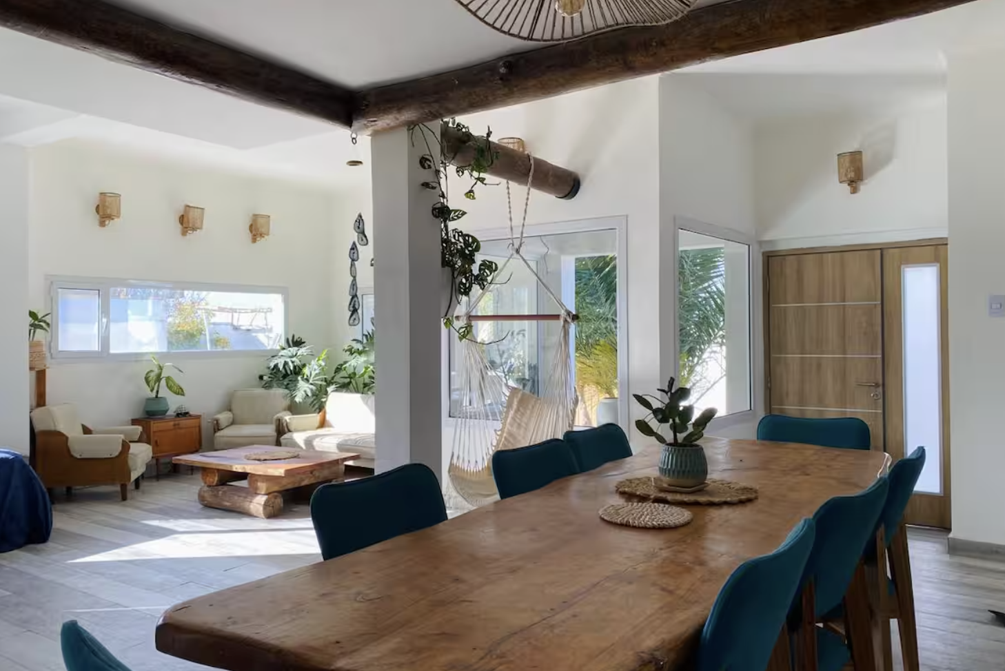 Open-concept living and dining room with wooden furniture, green plants, natural light from windows, and a hammock chair hanging from the ceiling.
