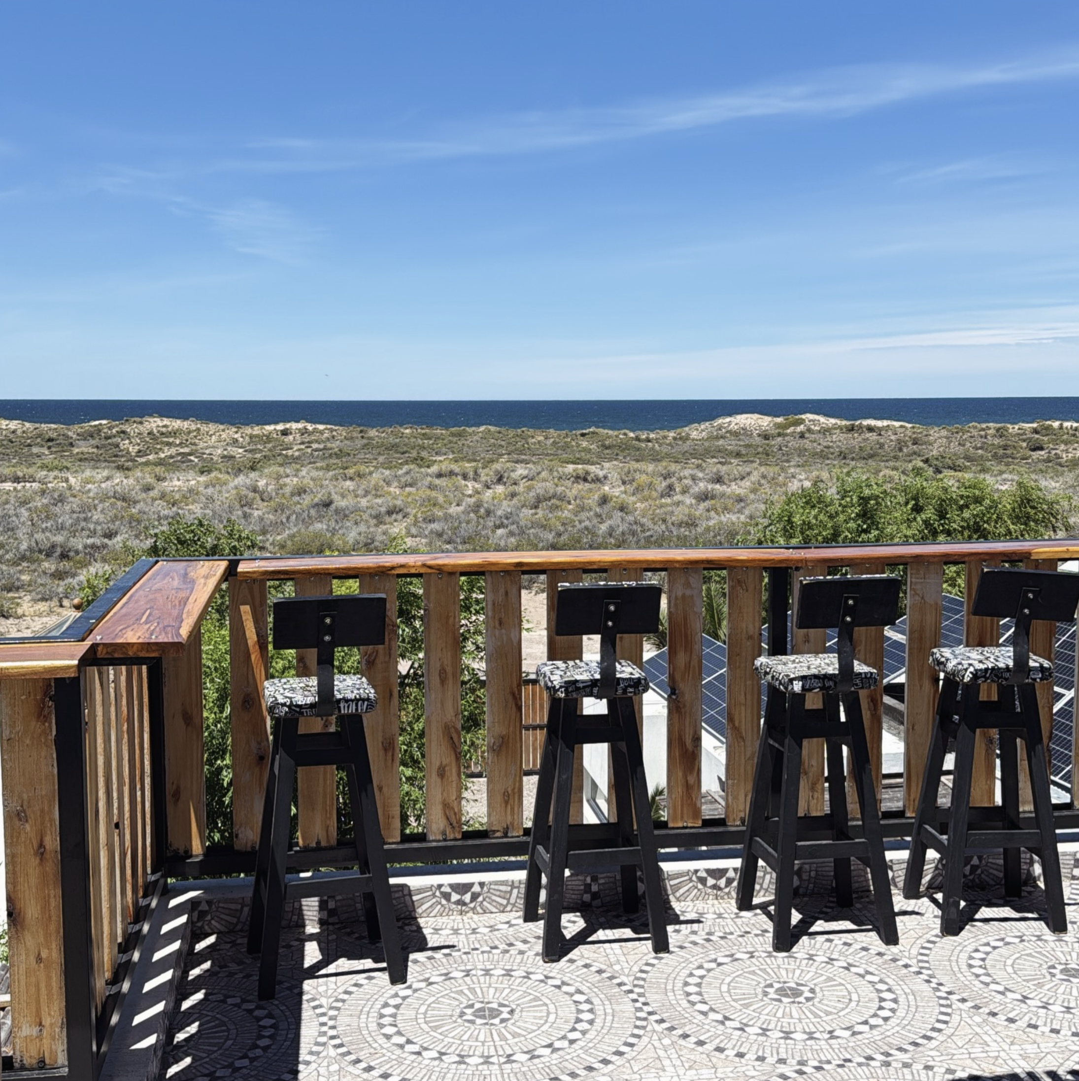 View from a wooden balcony overlooking a landscape with shrubs, a distant ocean, and a clear blue sky. Four black bar stools with patterned cushions are lined up against the railing.