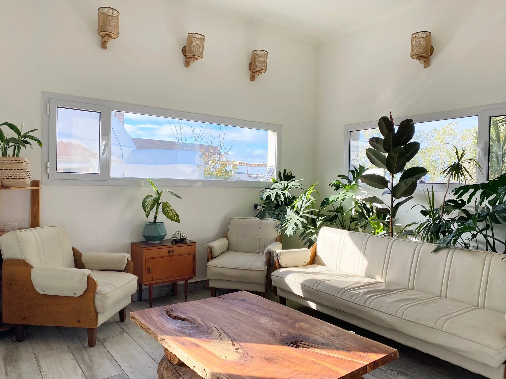 Living room with white sofas, a wooden coffee table, potted plants, large windows, and wall-mounted lamps.