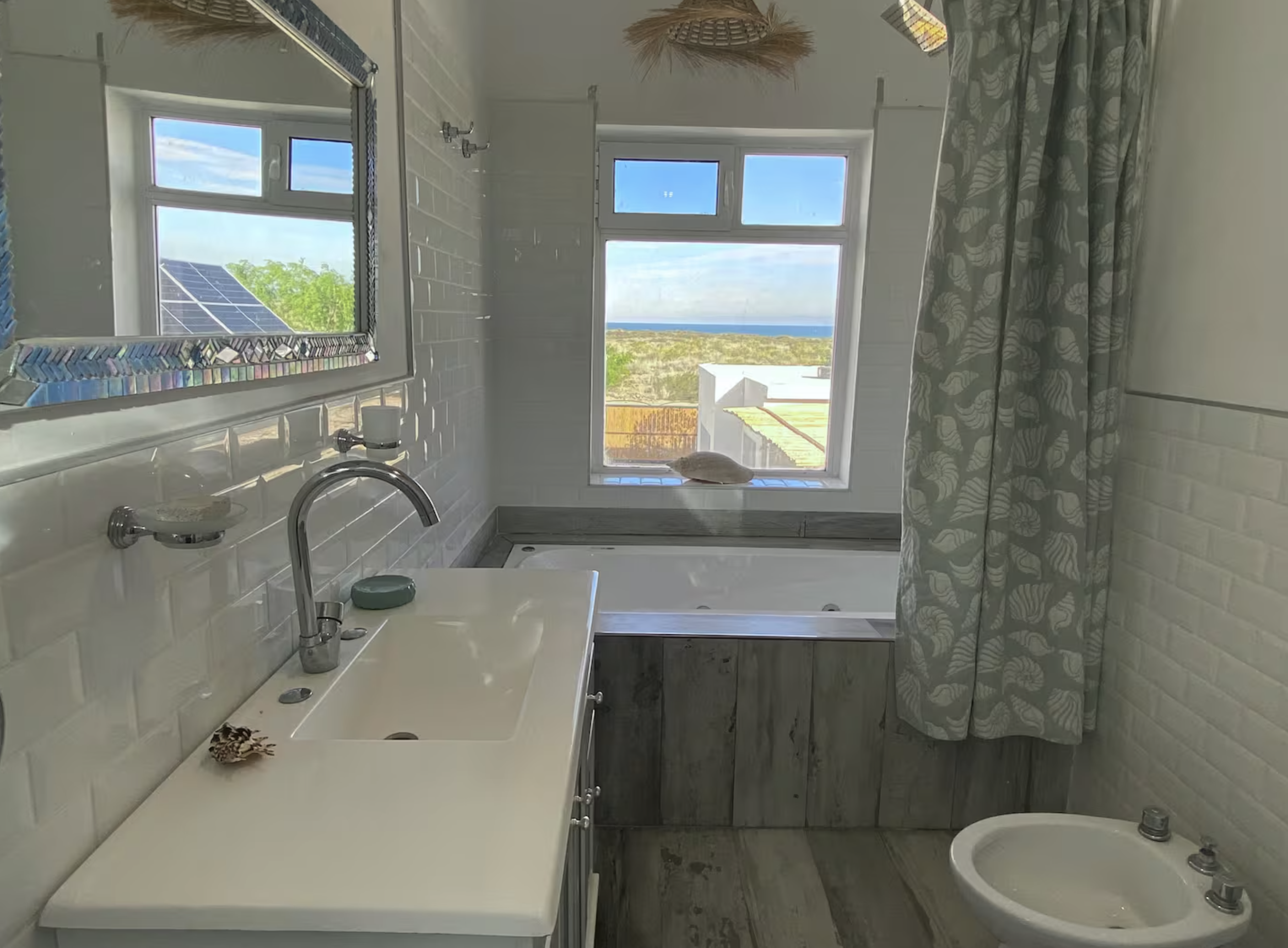 Bathroom with white brick wall, large window showing beach view, bathtub, sink, mirror, and shower curtain with a seashell pattern.