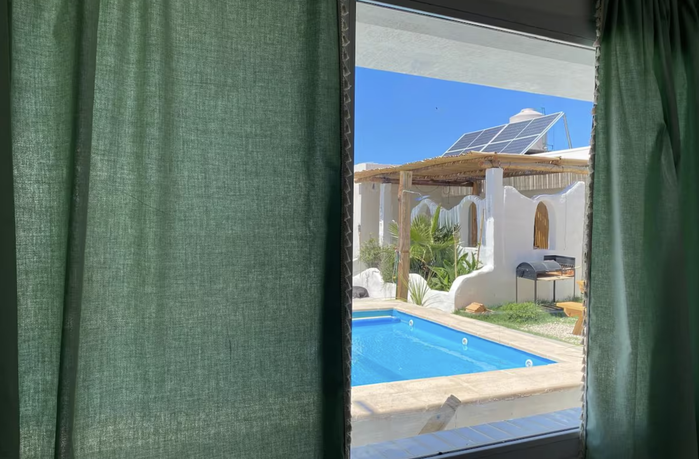 View of a backyard pool area through an open window with green curtains, showing a blue swimming pool, a small shaded structure, plants, and a building with solar panels on the roof under a clear blue sky.