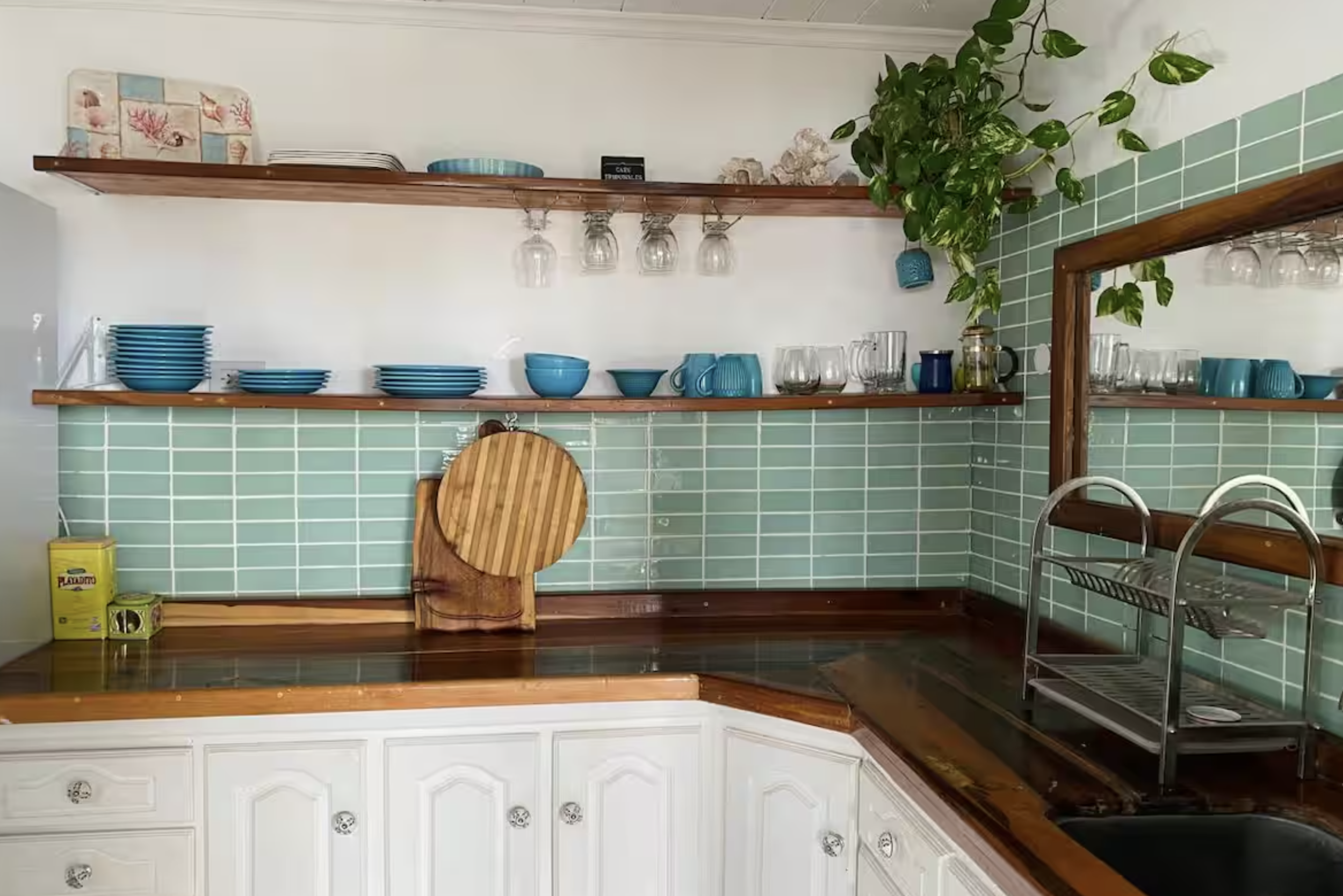Open kitchen with green tiled backsplash, white cabinets, and wooden countertops. Blue dishes and glasses on open shelving, a potted plant next to the mirror, and a wooden cutting board resting against the wall.