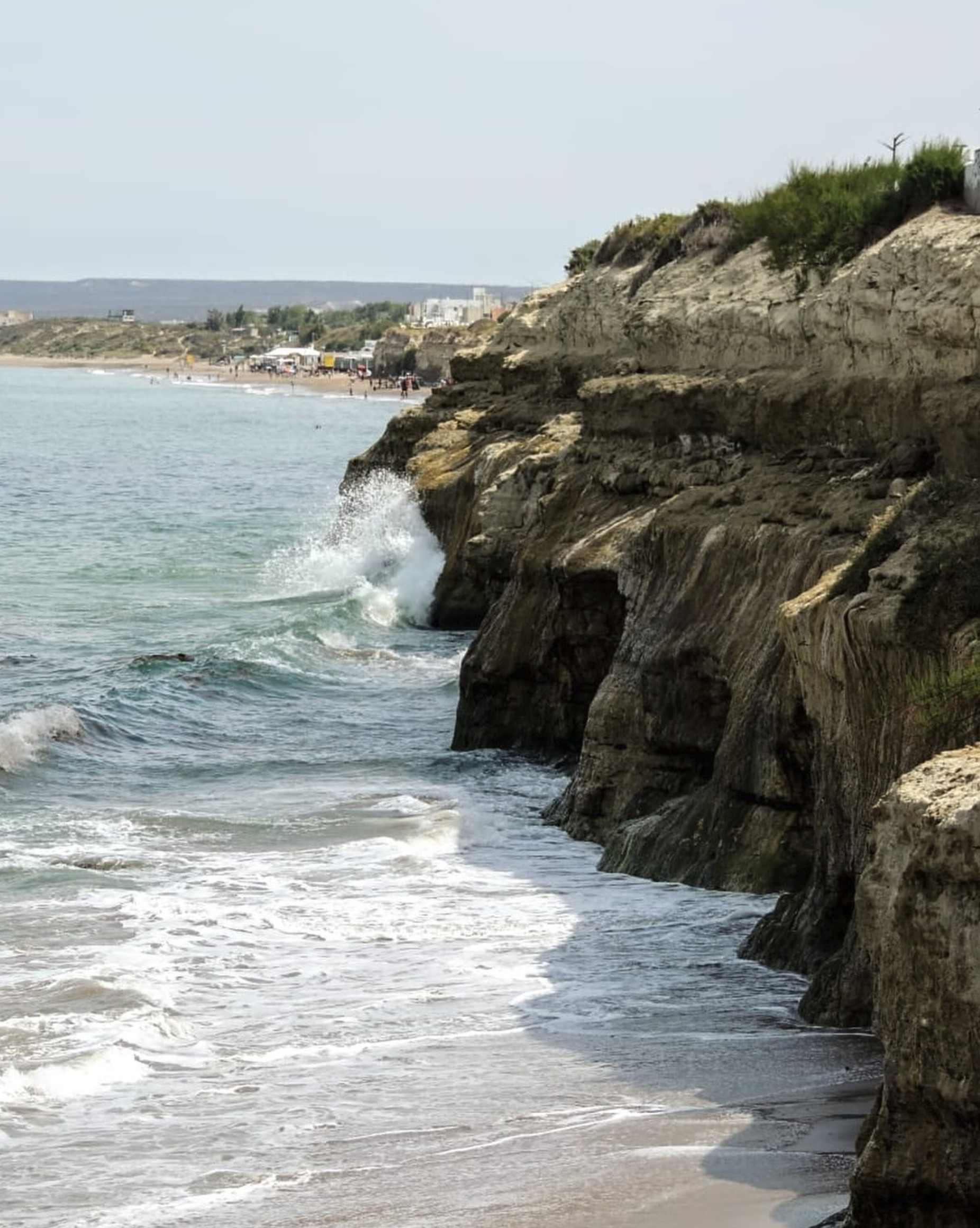 Cliffside view of a sandy beach with waves crashing against rock formations and a distant shoreline with people, vehicles, and buildings.