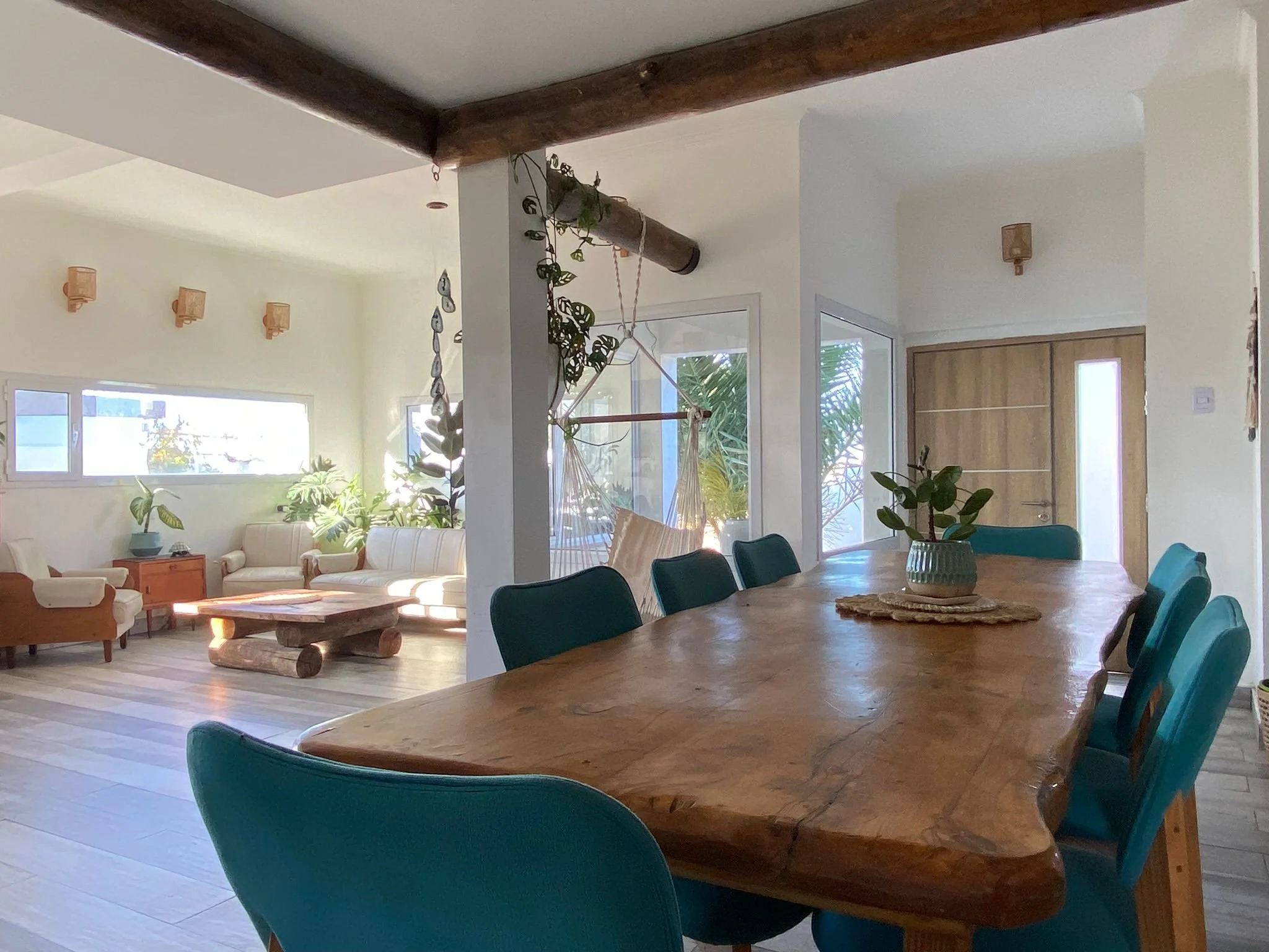 A bright, open living and dining room with large windows, a wooden dining table surrounded by teal chairs, and a cozy sitting area with white sofas and plants.