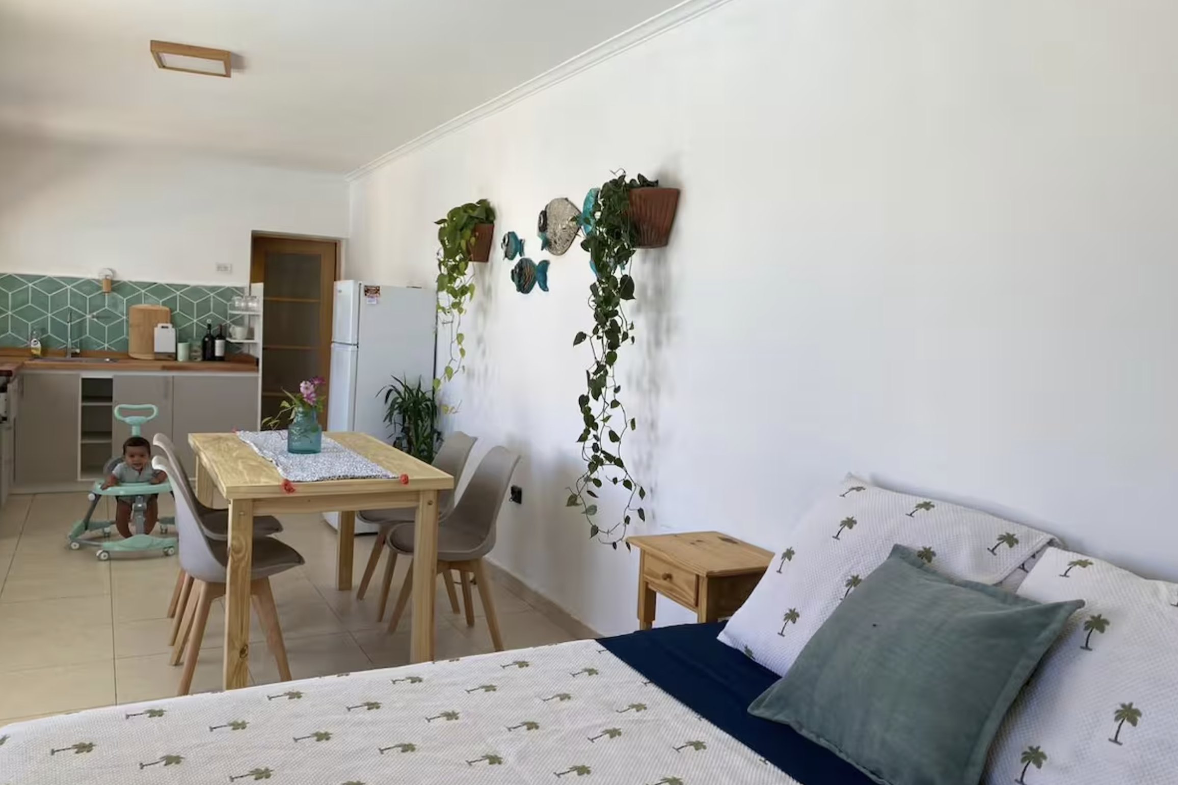 Open-plan living space with a bed, dining area, kitchen, and a child in a walker. Decor includes hanging plants, fish-shaped wall art, and a table centerpiece with flowers.