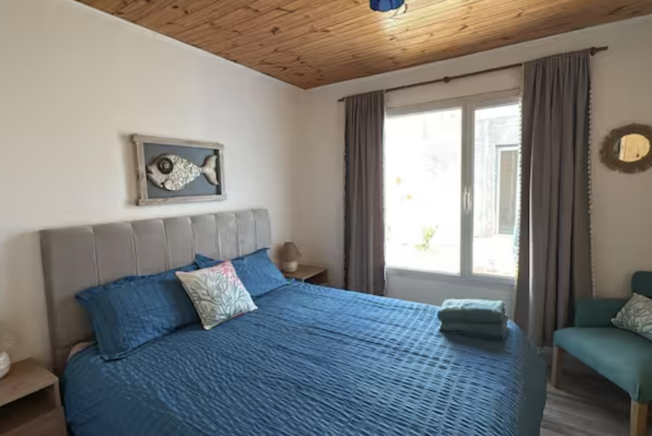 Bedroom with a bed covered in a blue quilt, a gray headboard, a pillow with a botanical pattern, and folded green towels on the bed. There are two nightstands with lamps, a teal chair, curtains, and a wooden ceiling.