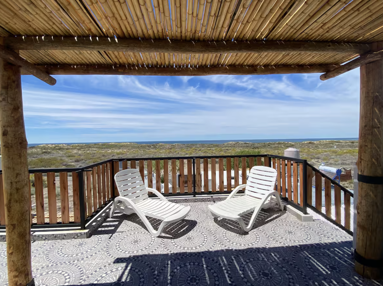 Beach view from a shaded wooden patio with two white lounge chairs and a wooden railing, overlooking sand dunes and the ocean under a partly cloudy sky.