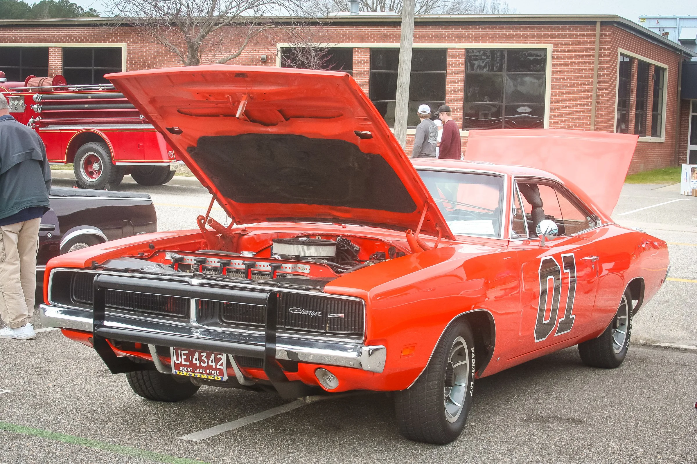 Red vintage Dodge Charger muscle car with its hood open, parked at a car show on a street in front of a brick building. Several people are walking around and viewing other cars.