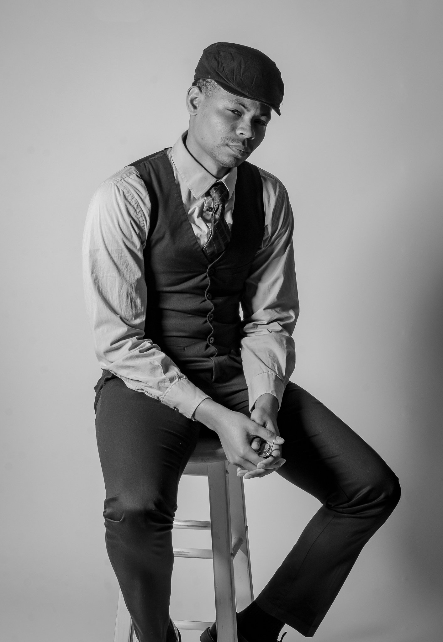A man sitting on a stool, dressed in a button-up shirt, tie, vest, and cap, with a serious expression, in black and white.