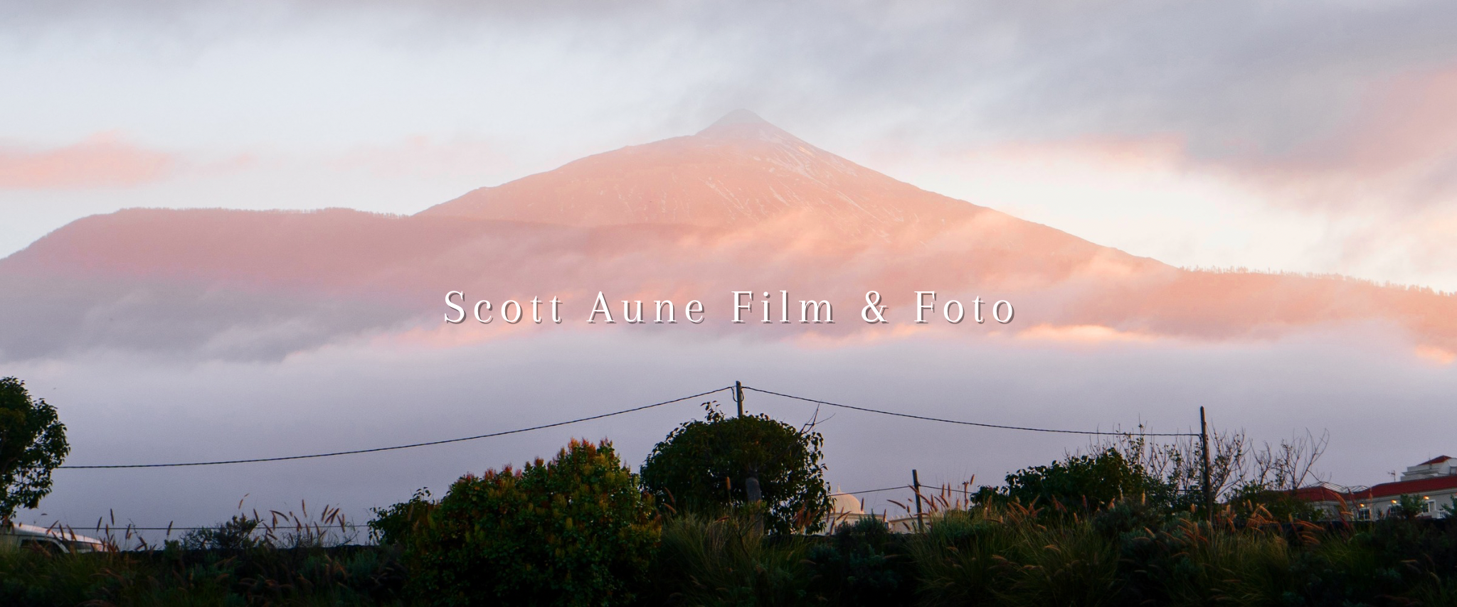 Fjell med tåke og skyer, med hus og trær i forgrunnen, soloppgang eller solnedgang.
