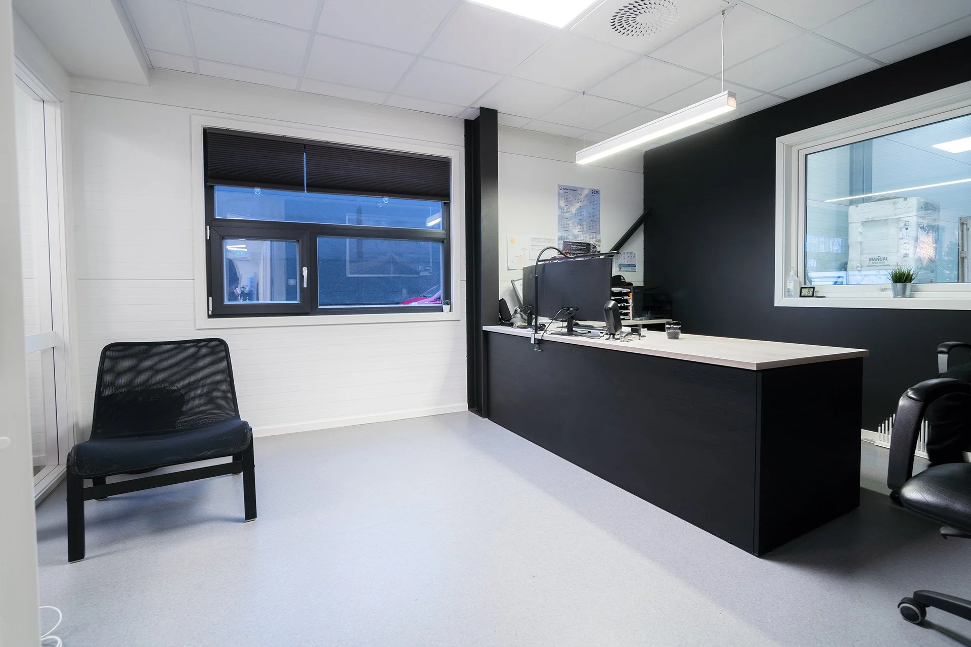 Et kontor med hvite og svarte vegger, to vinduer, en svart og en hvit stol, et svart skrivebord med datamaskin og utstyr, og et opplyst lys på taket.