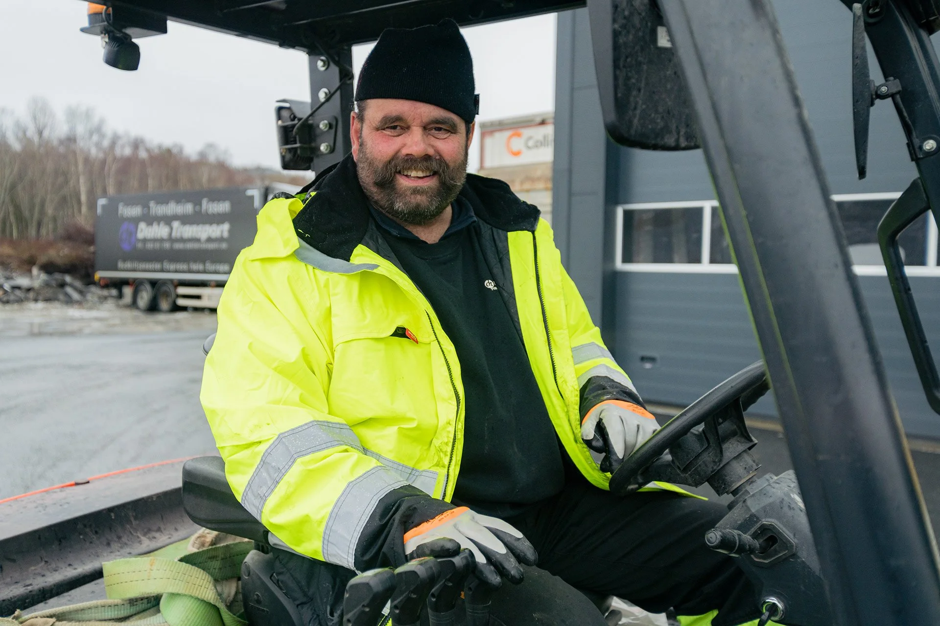 En mann i høy synlighetjakke og svart lue kjører en gravemaskin utendørs.