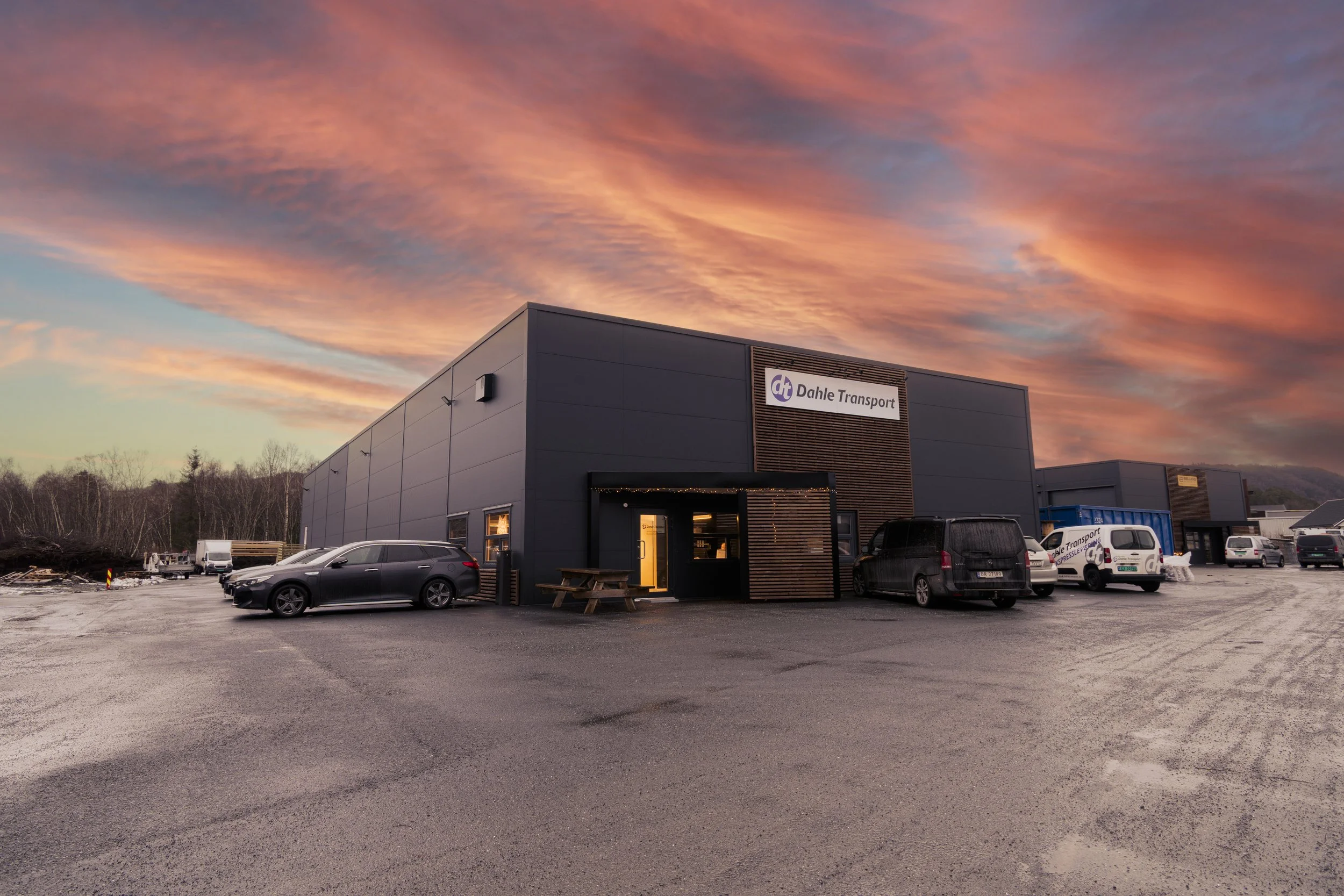 En modern industribygning med en parkeringsplass foran, flere biler parkert, under en fargerik solnedgang. Skiltet viser 'Dahle Transport'.