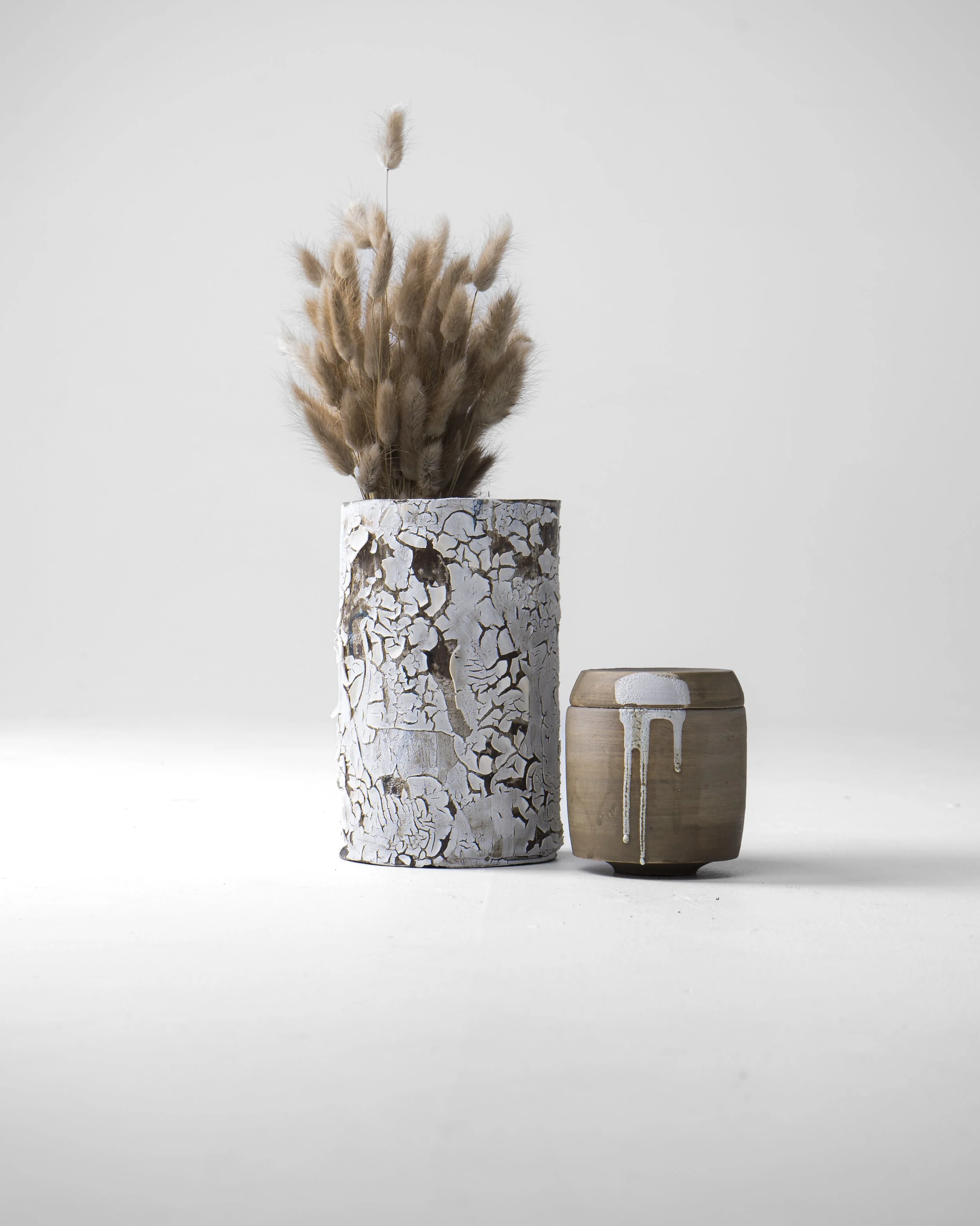 To keramikkrukker, en med hvit, sprukket maling og en med brun overflate, oppstilt mot en lys bakgrunn. Den ene krukken har tørkede, bustete planter oppi. Kathrine Berg keramik