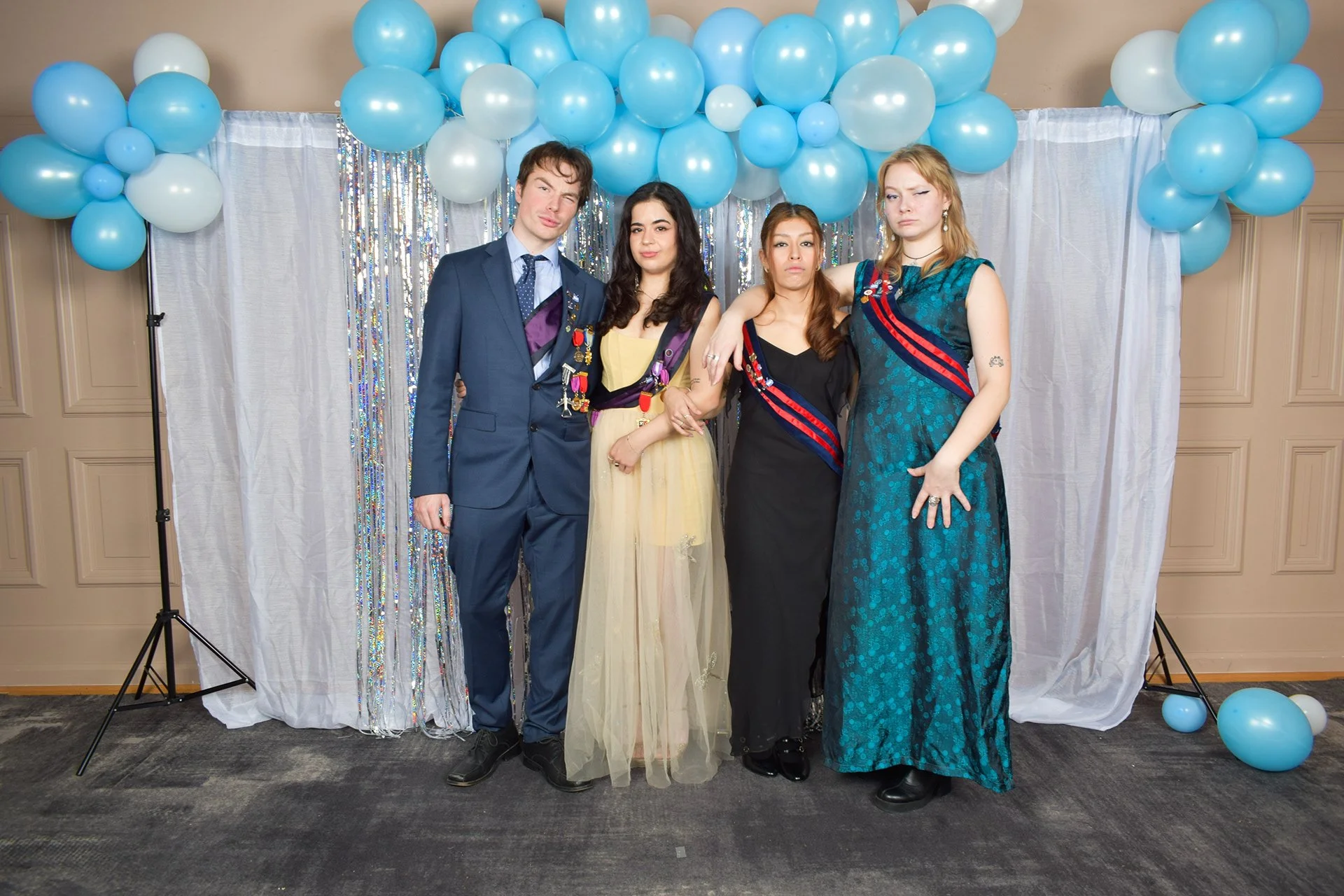 Fire personer kledd i formelle antrekk, står foran dekorasjoner med blå ballonger og hvite gardiner.