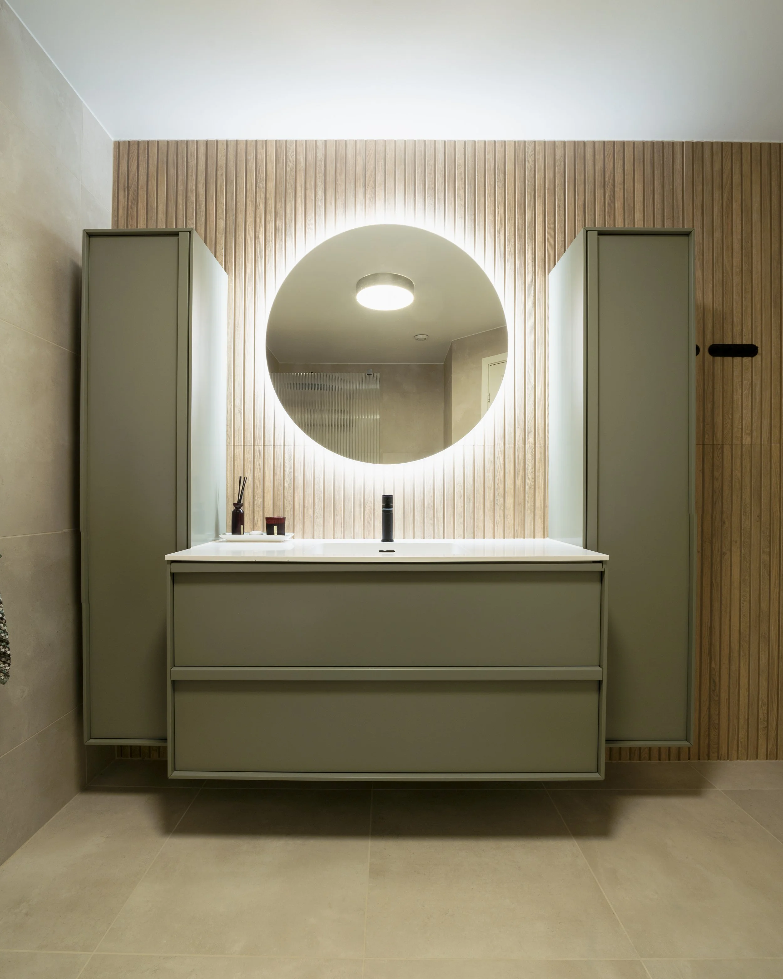 Moderne bademøbel med stor rund speil med belysning, to skuffer, og utstyr som tannbørsteholder og liten beholder, foran en vegg med trepanel.