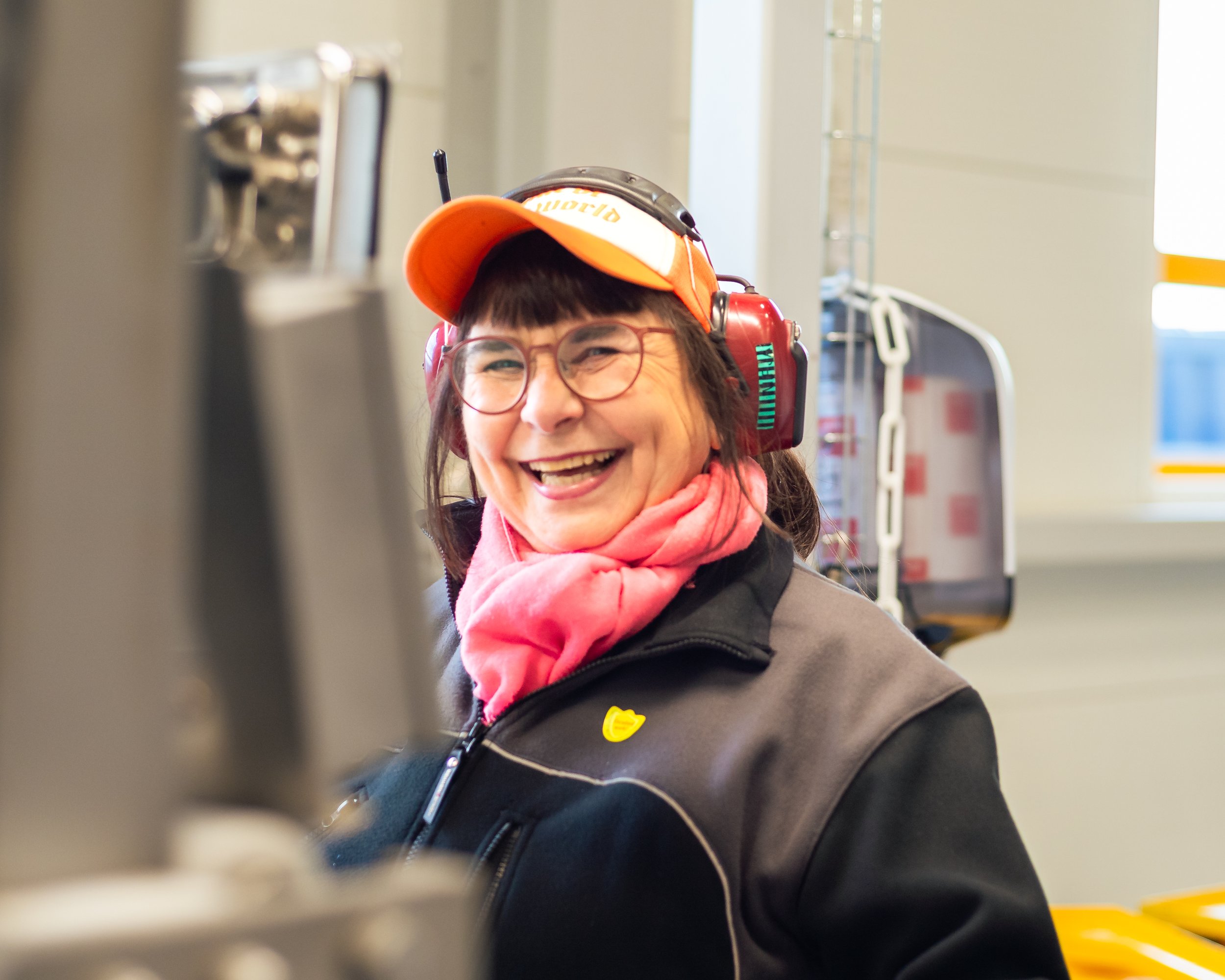 En kvinne smiler og ler, iført hjelm, briller, oransje caps og rosa skjerf, i en industri eller arbeidsmiljø.