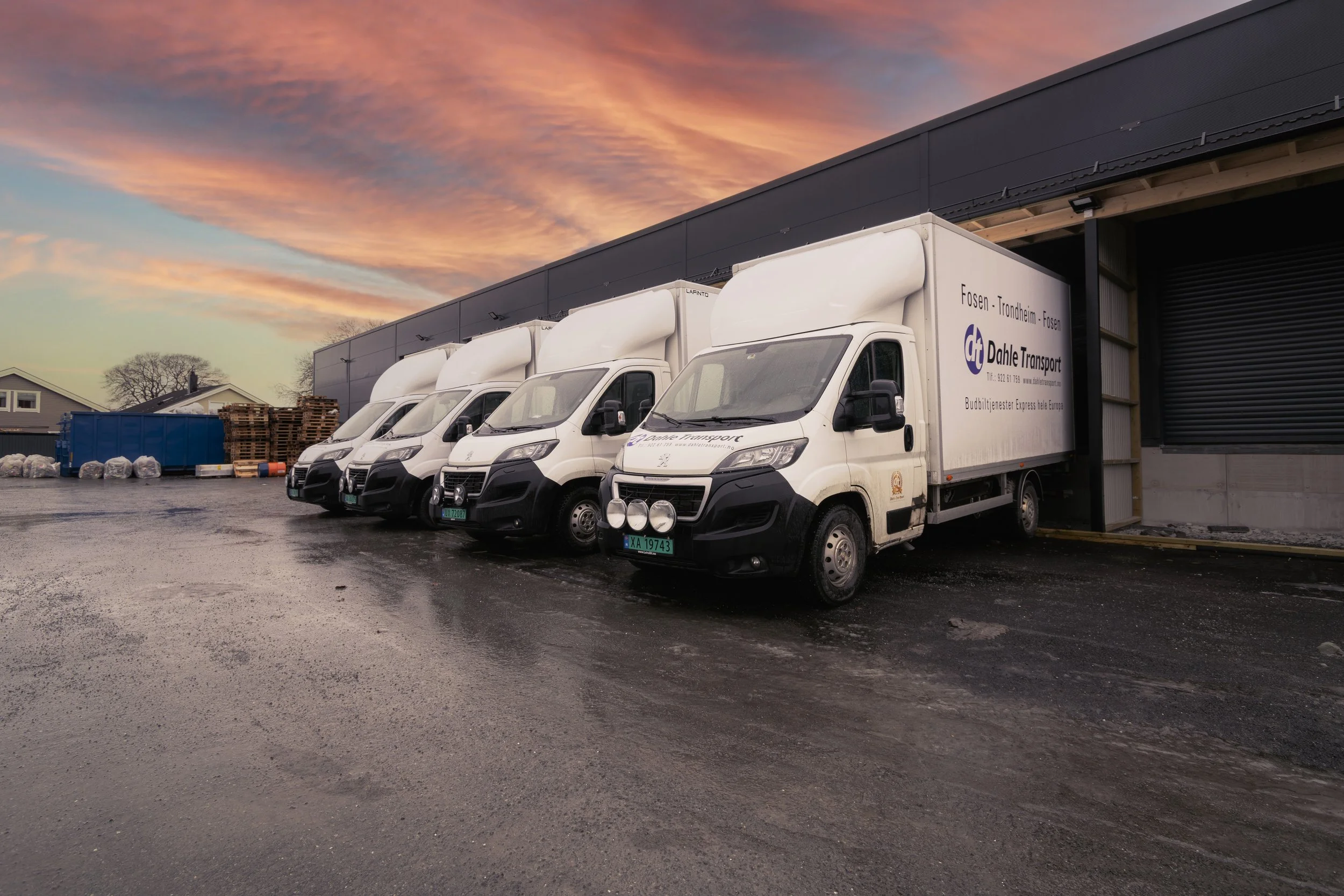 Fire hvit varebiler parkert utenfor en lagerbygning mot solnedgang, med en blå container og noen paller i bakgrunnen.