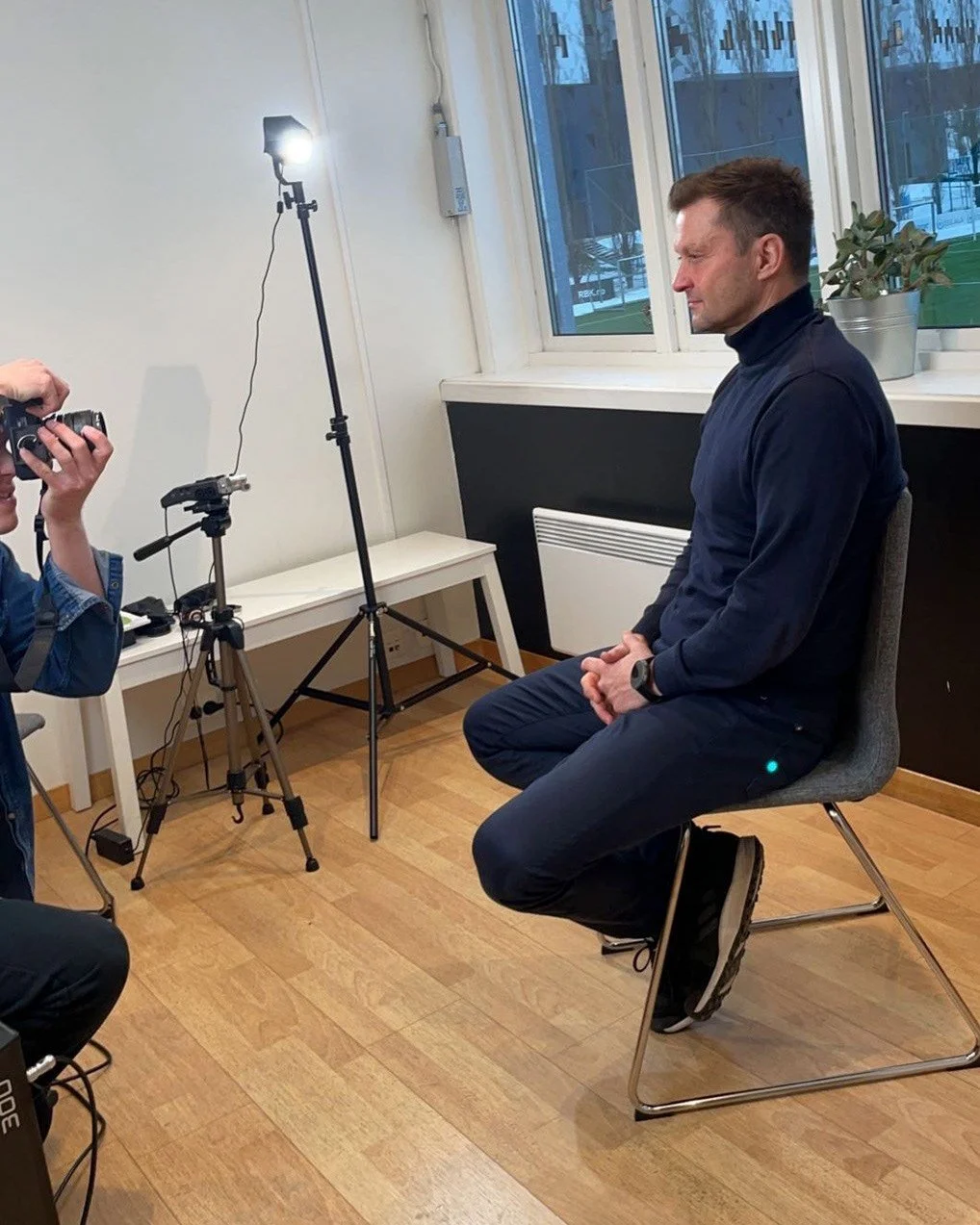 En mann med kort hår og sportsklær sitter på en stol under en fotoshoot eller intervju, med et kamera fotografen holder opp. Det er en lite rom med vindu og innendørs planter.