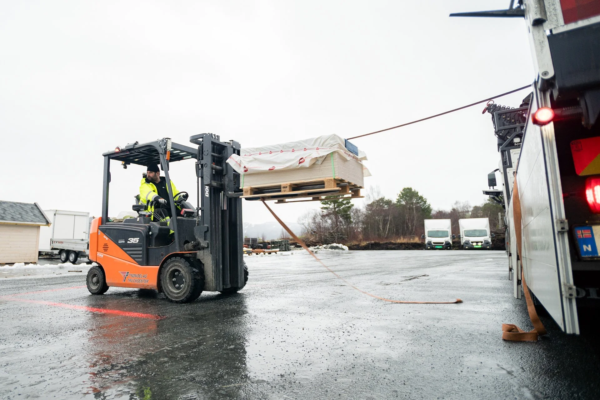 En person kjører gaffeltruck med en pall lastet med materialer på en våt asfaltflate, omgitt av lastebiler og trær.