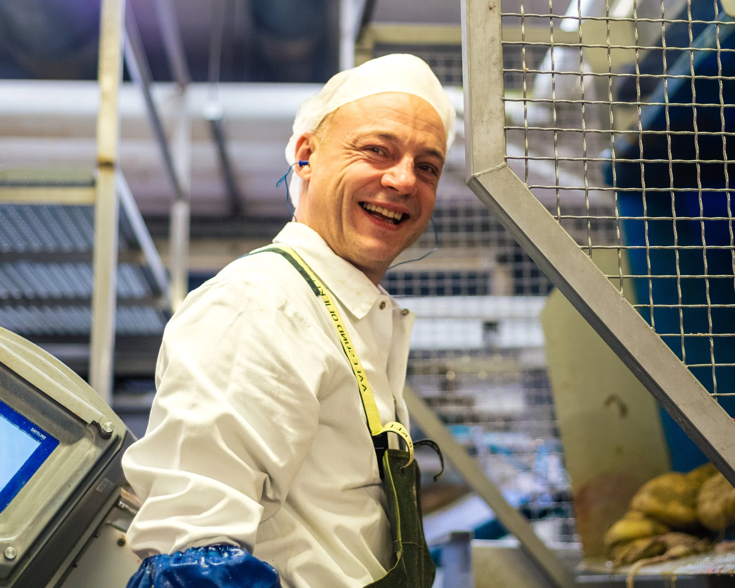 En mann smiler på en fabrikk eller arbeidsplass, iført hvitt arbeidstøy, blå hansker og med headset, omgitt av metallhyller og emballerte produkter.