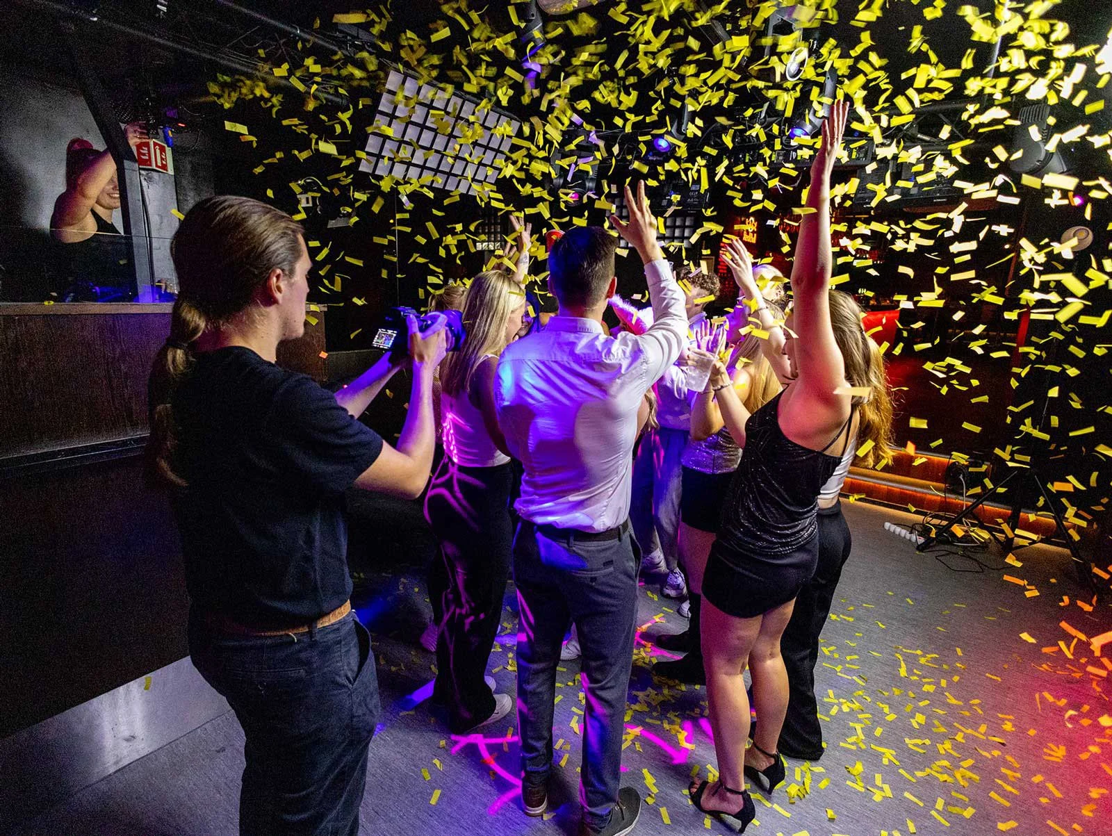 Personer danser og feirer på en klubb eller et arrangement med gul konfetti som faller ned. Noen tar bilder, og DJ-en er synlig i bakgrunnen.