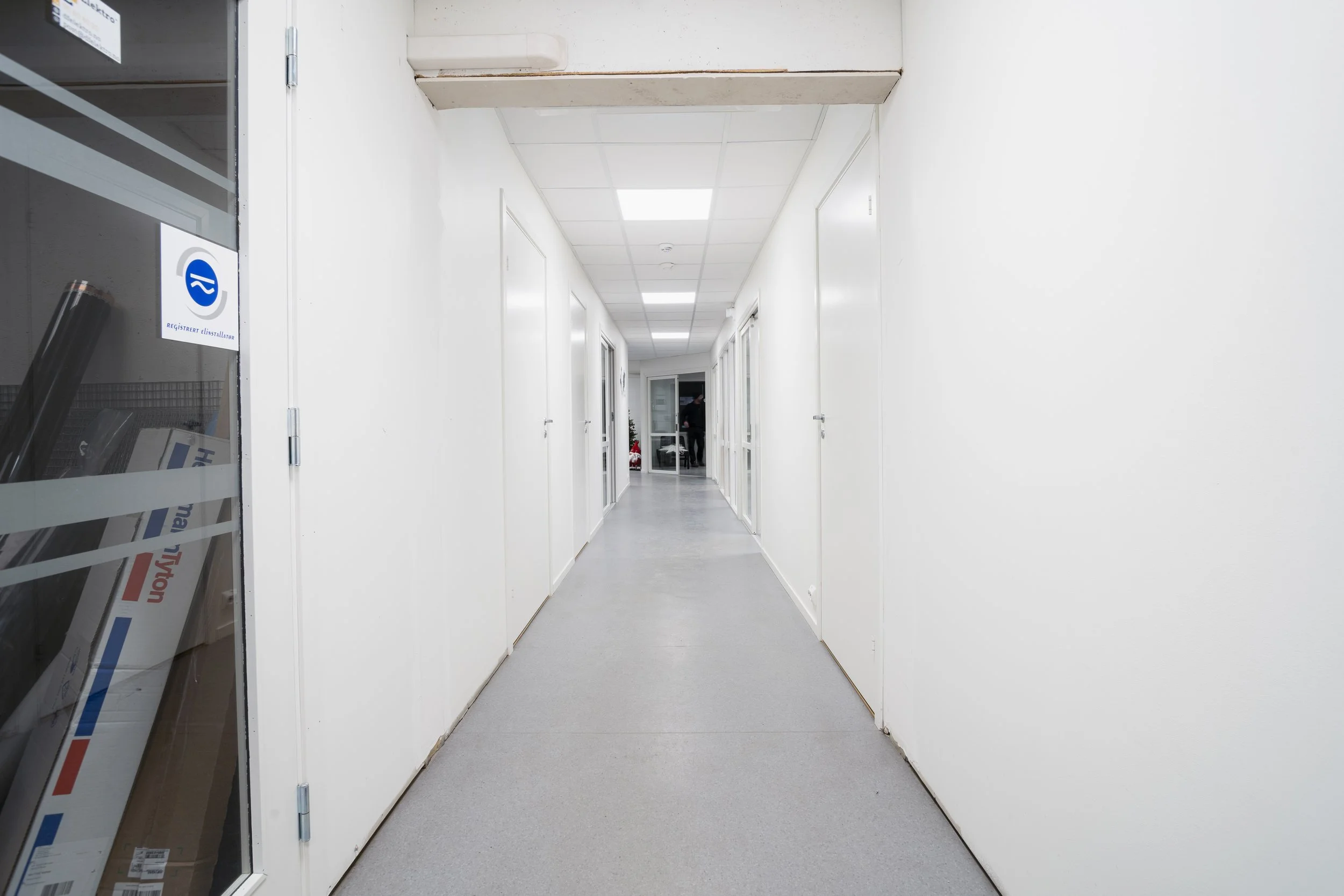 Et langt, hvitt, innendørs kontorlignende korridor med dører på begge sider og et vindu med fargerike akrylbilder til venstre. I bakgrunnen står det en person nær en glassdør.