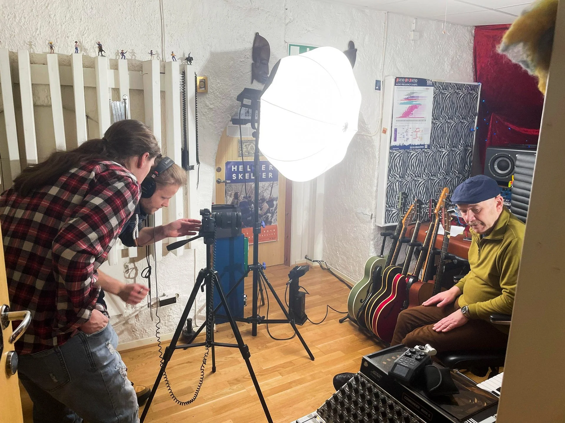 To personer som filmer i et musikkstudio, med gitarer stående mot veggen, et stort lys og lydutstyr.