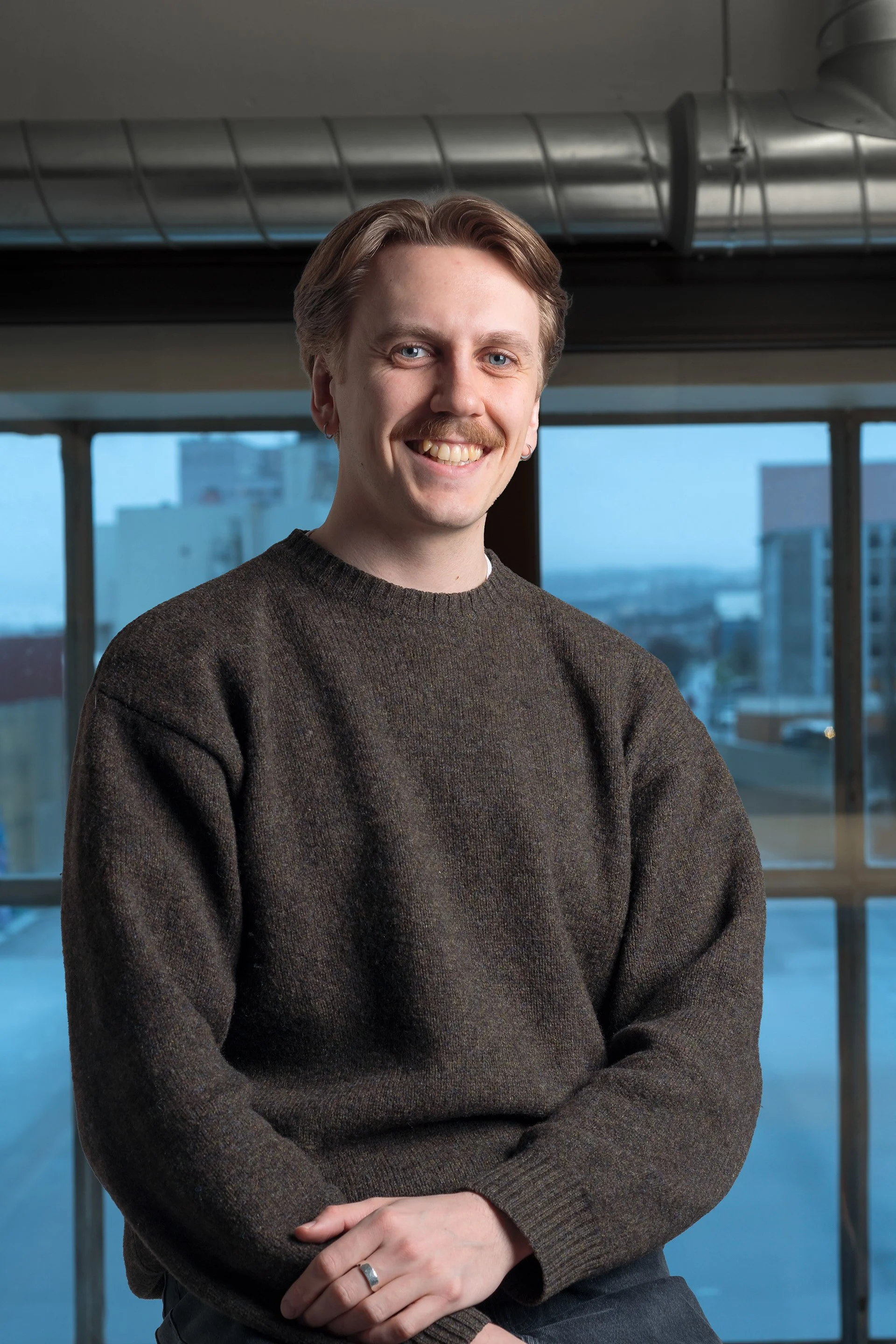Et smilende mennske portrett foran store vinduer med bylandskap i bakgrunnen.