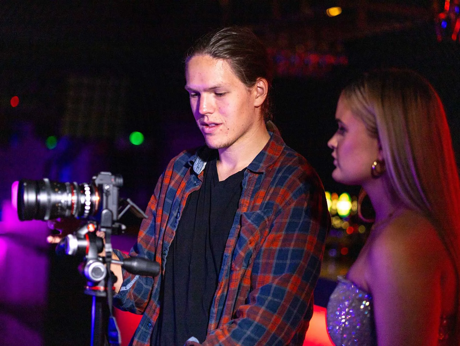 En mann og en kvinne ser på en kamerastabilisator i en nattklubb med fargerike lys i bakgrunnen.