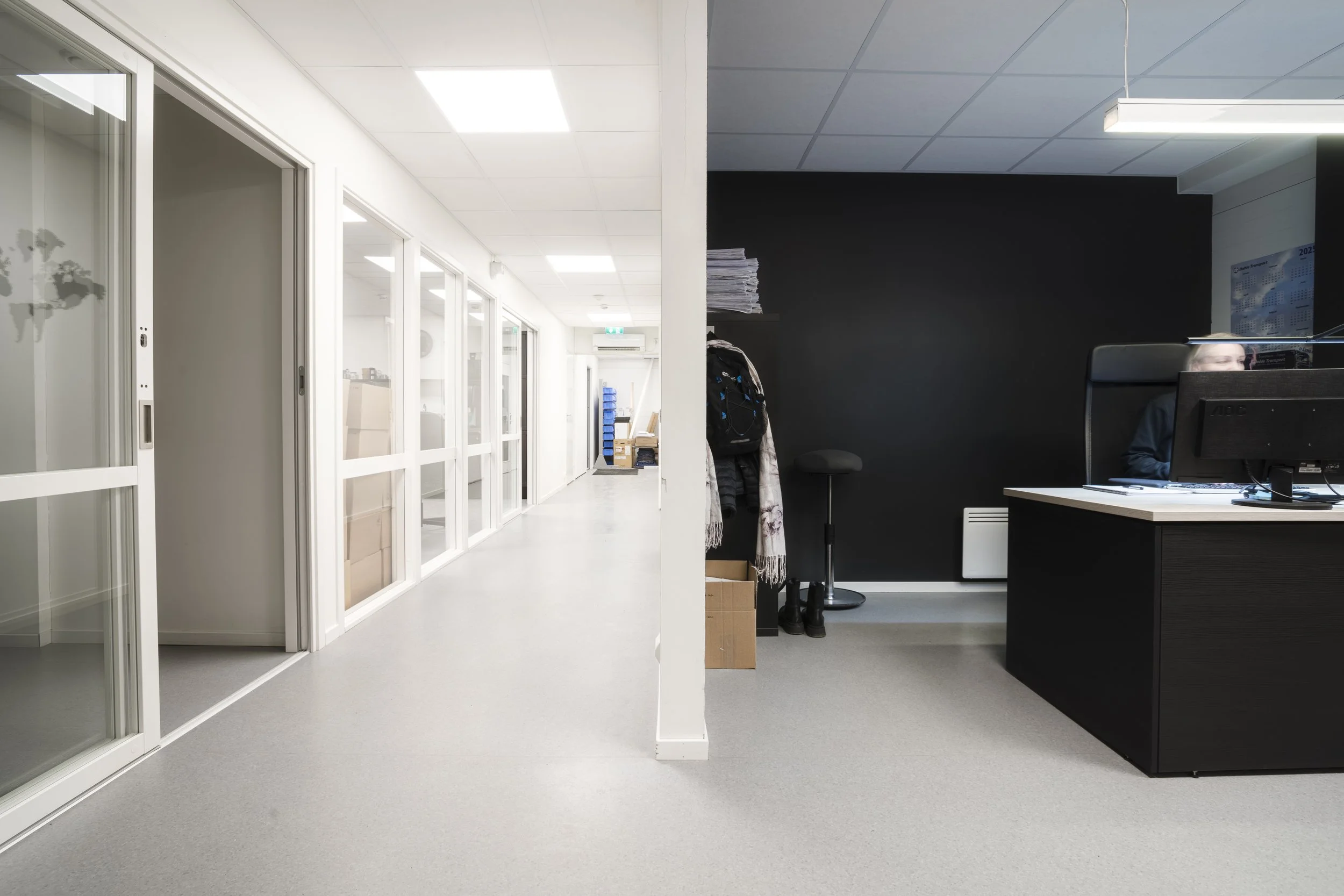 En tom kontorrekke med glassvegger på venstre side og en resepsjonsskranke på høyre side, med en svart vegg og et søppelstativ i bakgrunnen.