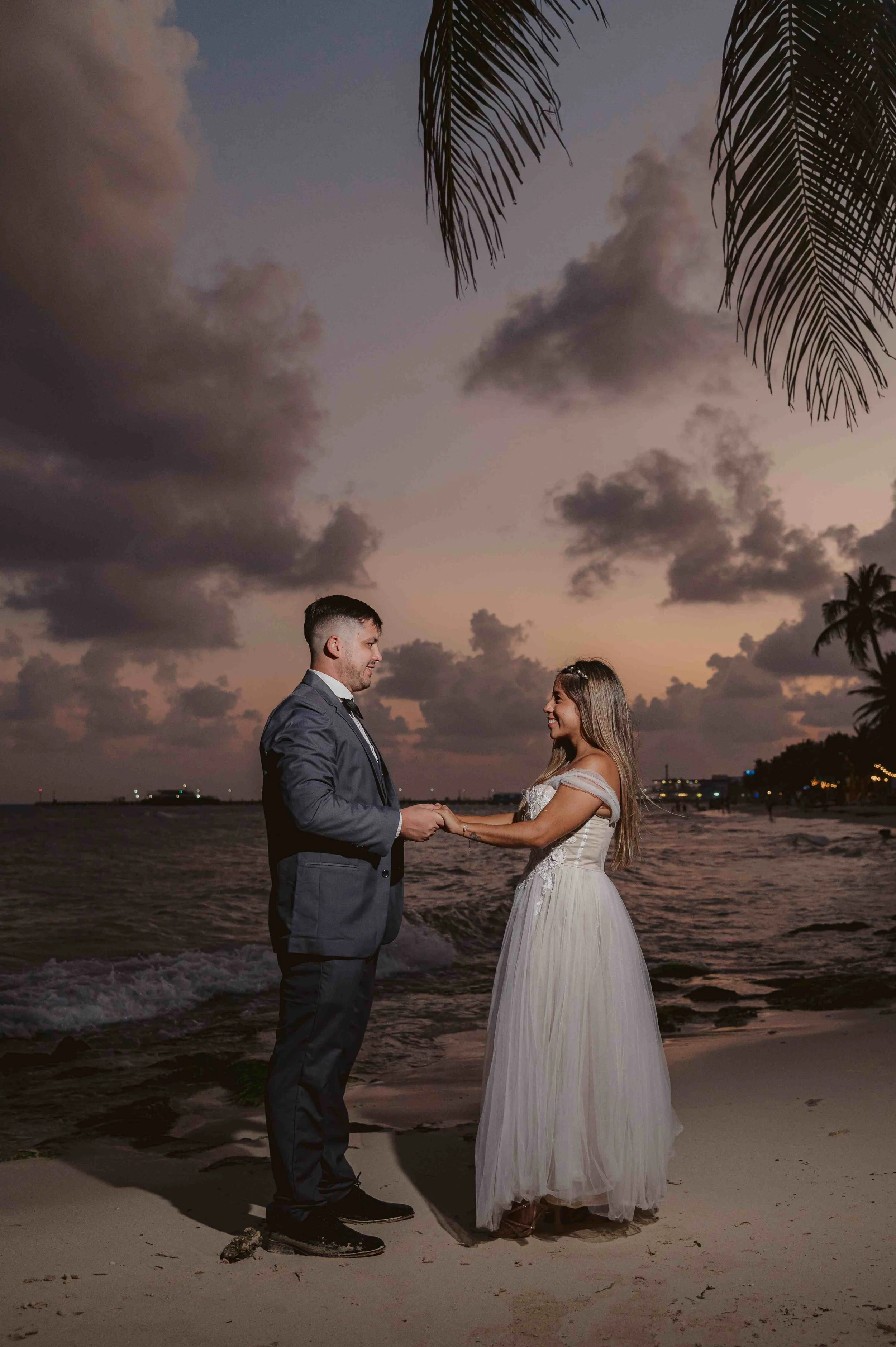 A couple, dressed in wedding attire, holding hands and smiling at each other on a beach at sunset with clouds and palm trees in the background.