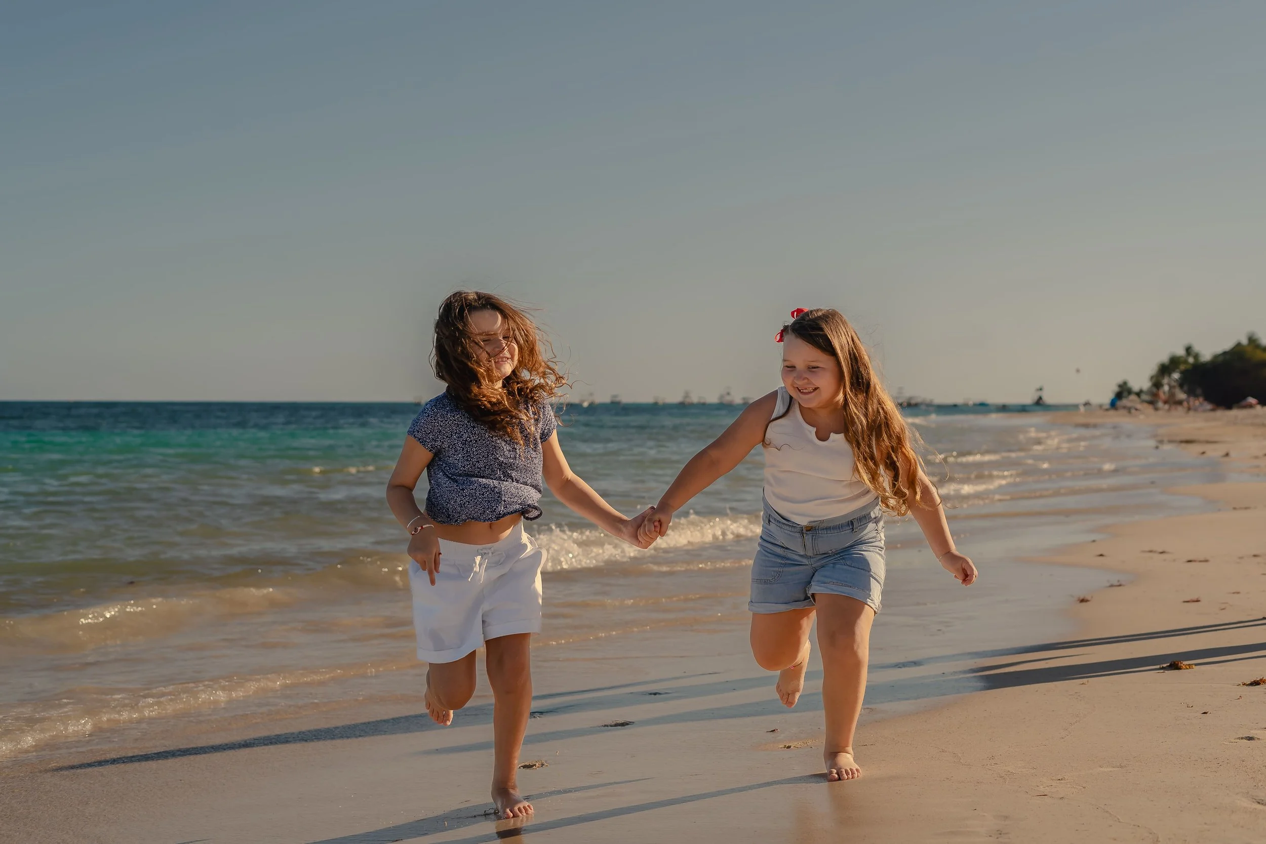 Two young girls running and holding hands on the beach near the water, smiling, with a clear sky and distant shoreline in the background.