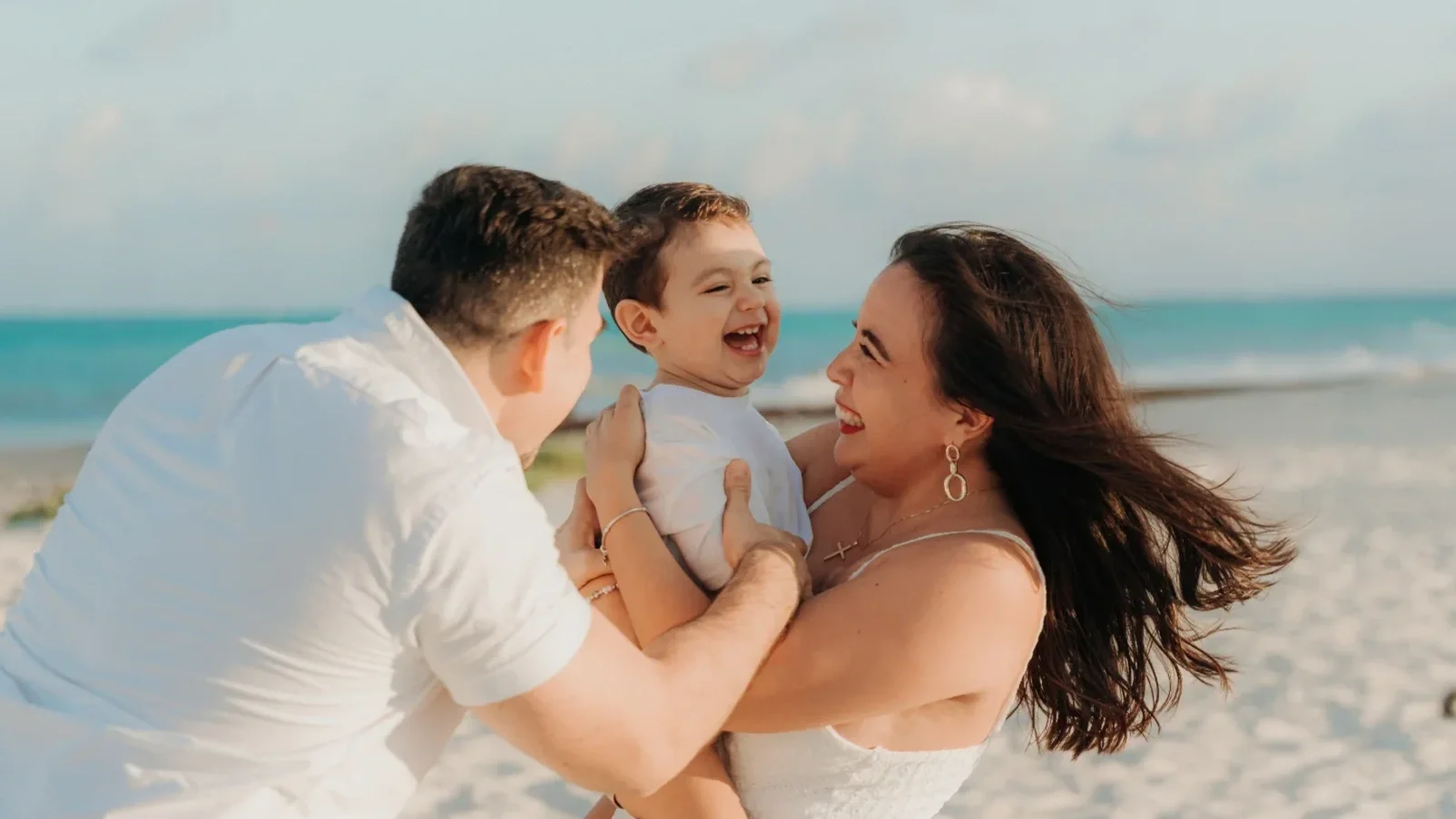 A family of three enjoying a playful moment at the beach, with the mother holding their young son and the father leaning in, all smiling and laughing.