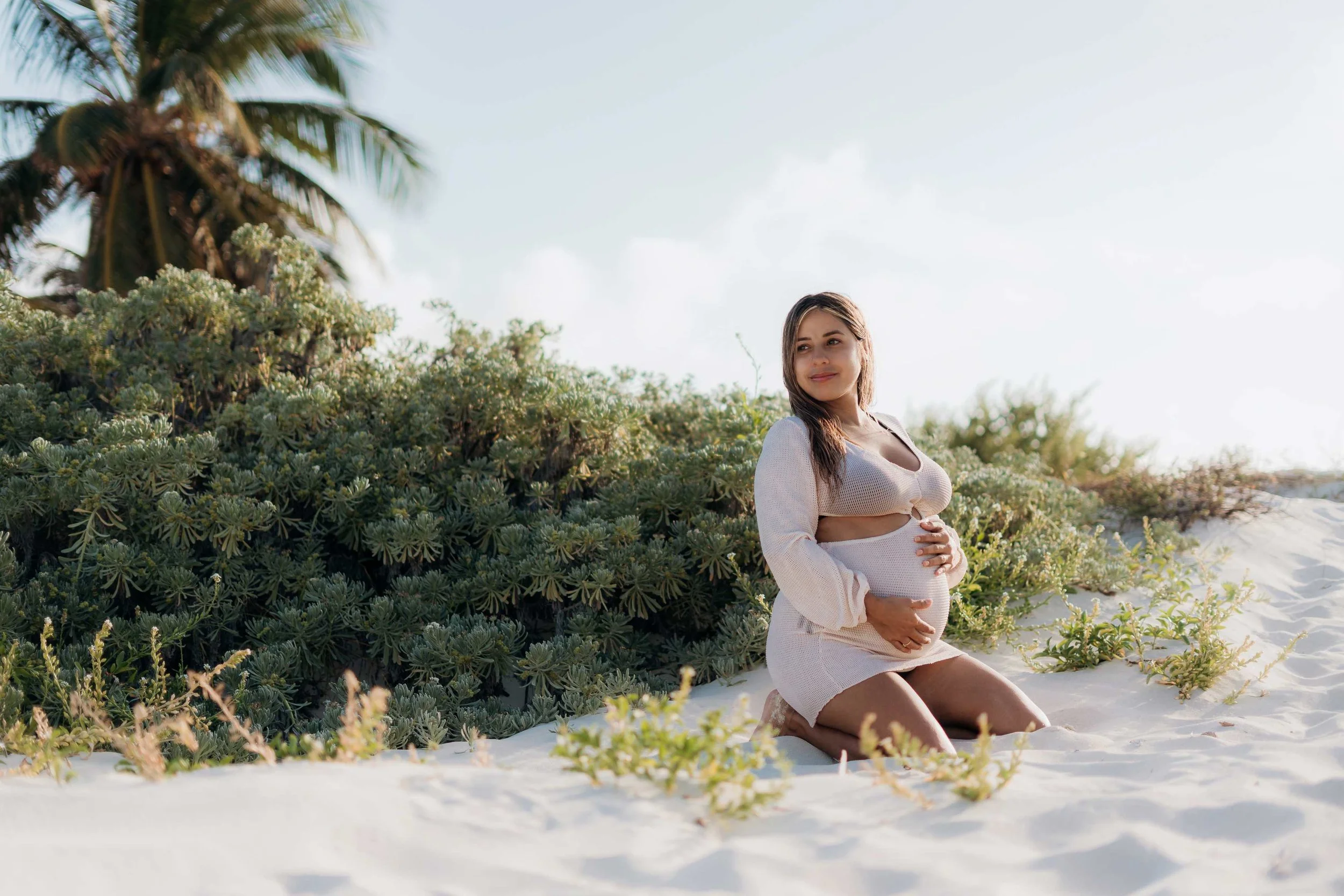 Pregnant woman on a Cancun beach kneeling on white sand, holding her belly, with lush green bushes and palm trees in the background, under a partly cloudy sky.