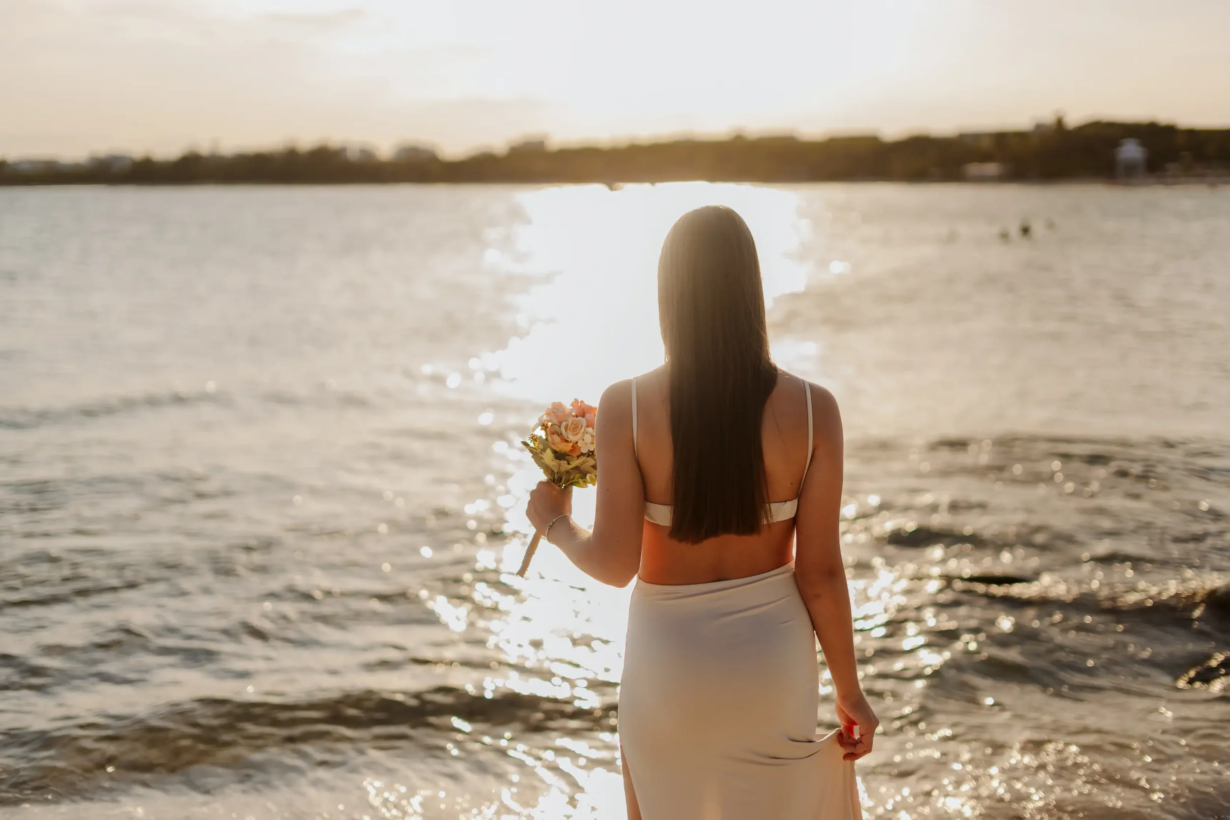 A woman with long dark hair in a white dress stands on the beach facing the sunset over the water, holding a bouquet of flowers.