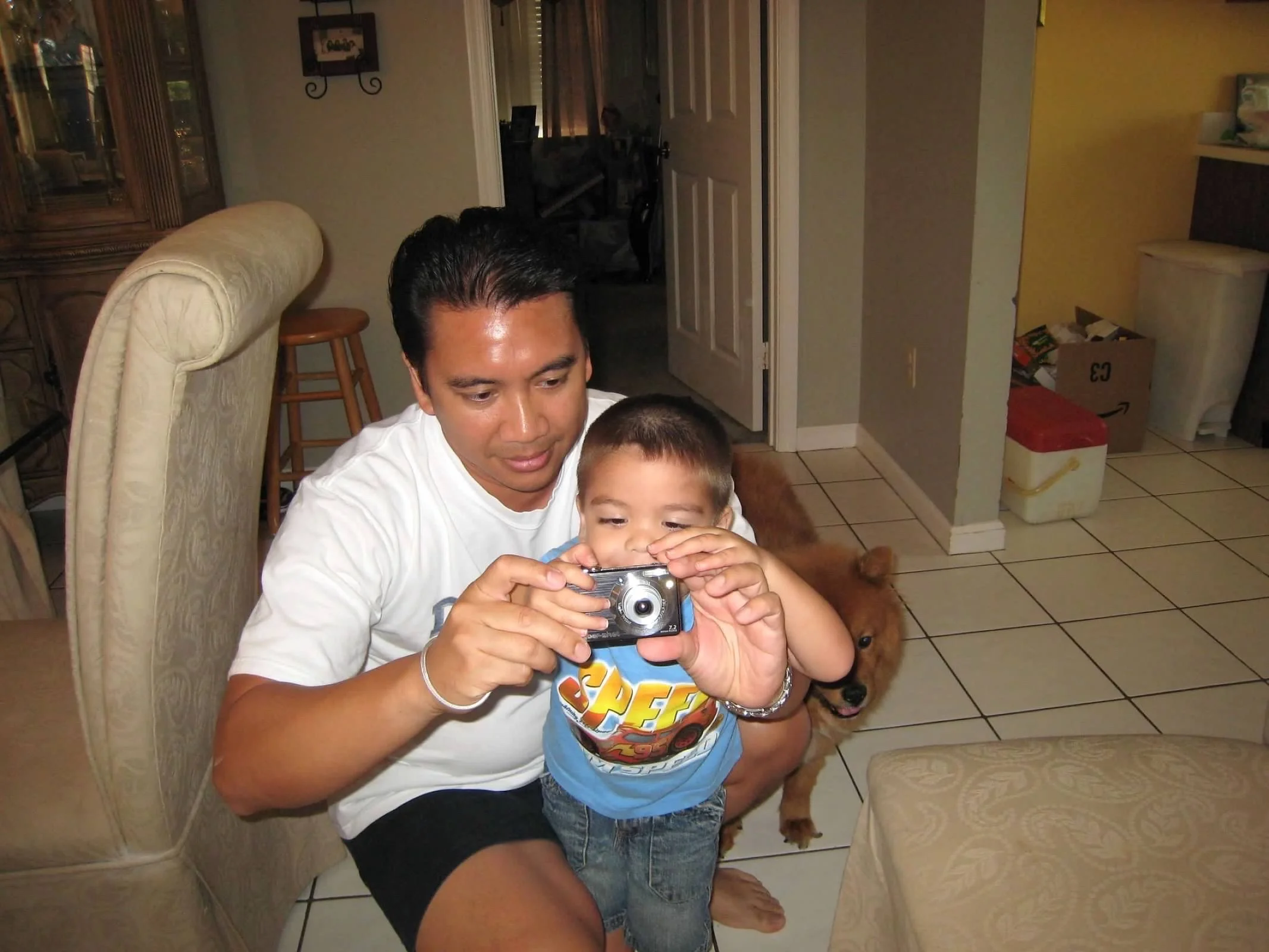 A man and a young boy are taking a selfie with a camera inside a living room. There is a golden retriever behind them. The man has dark hair and is wearing a white shirt, and the boy is wearing a blue shirt with a superhero logo.