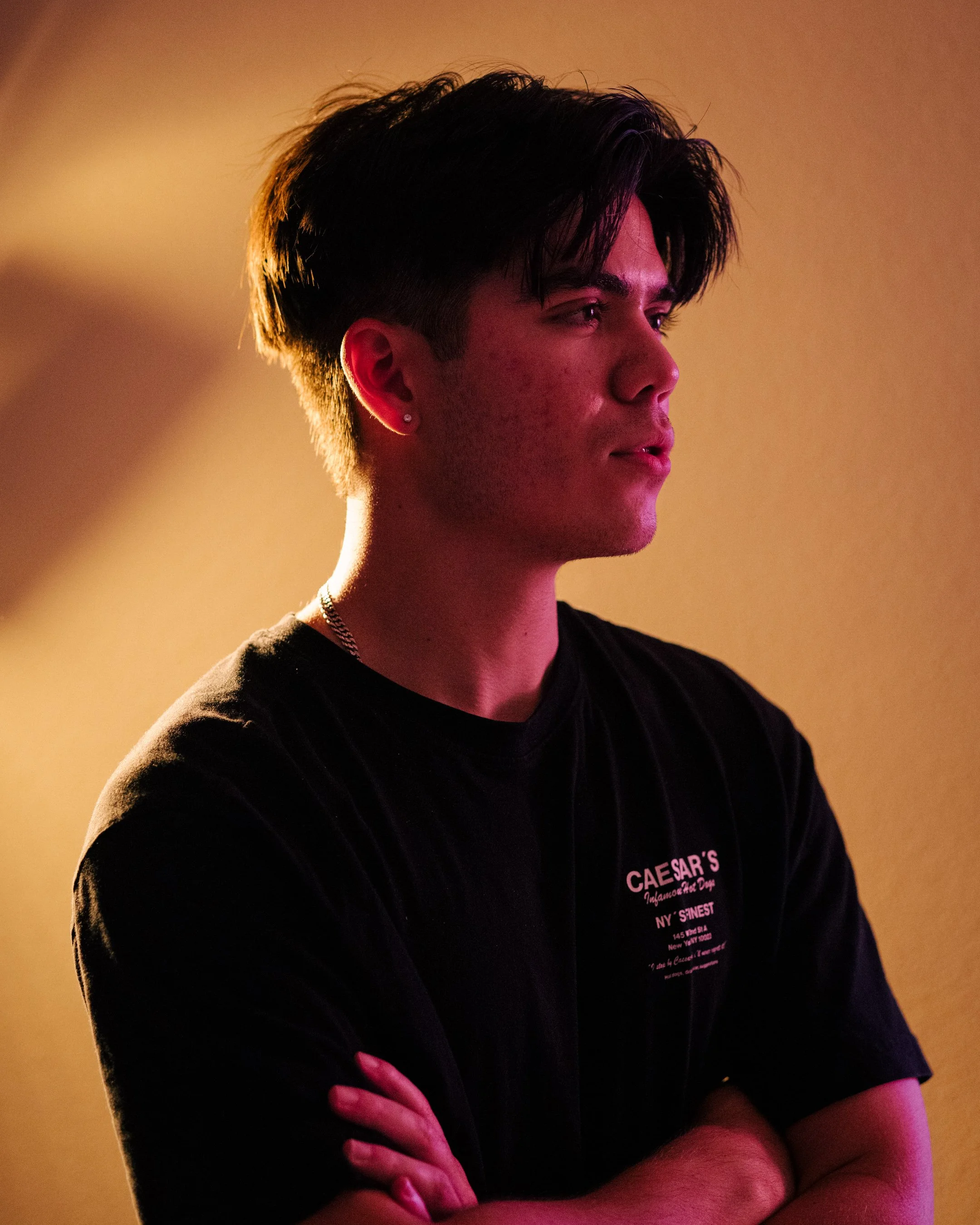 A young man with dark hair, wearing a black t-shirt, standing against a beige background with his arms crossed, looking to the side.
