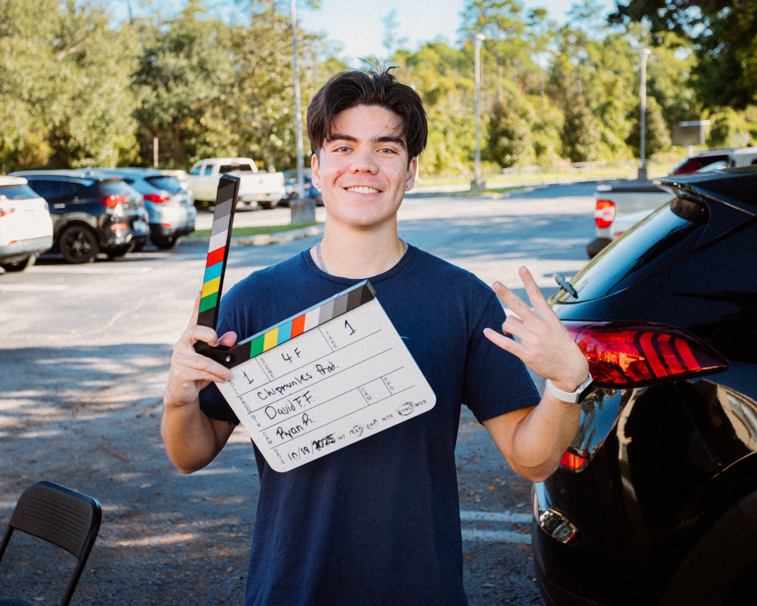 A young man holding a film slate in a parking lot, smiling and making a peace sign with his right hand, standing next to a black vehicle with a background of parked cars and trees.