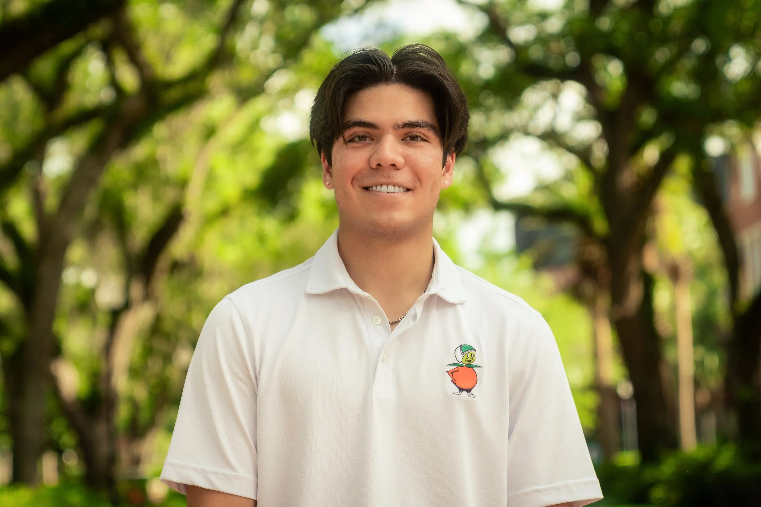A young man with dark hair smiling outdoors, wearing a white polo shirt with a colorful duck logo on the left chest area, and standing in front of green trees.